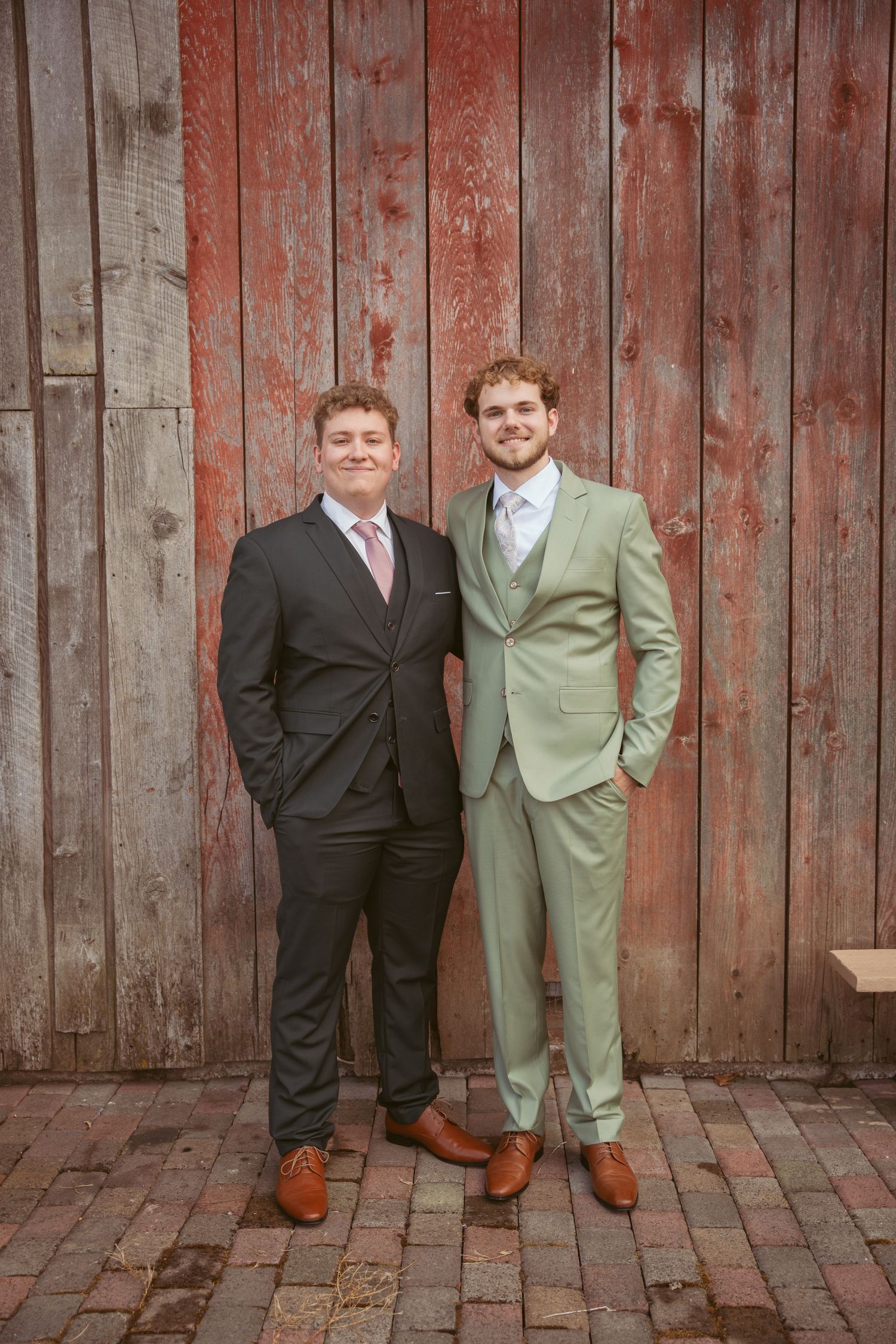 Groomsmen in formal attire stand together against weathered barn siding for wedding photos.