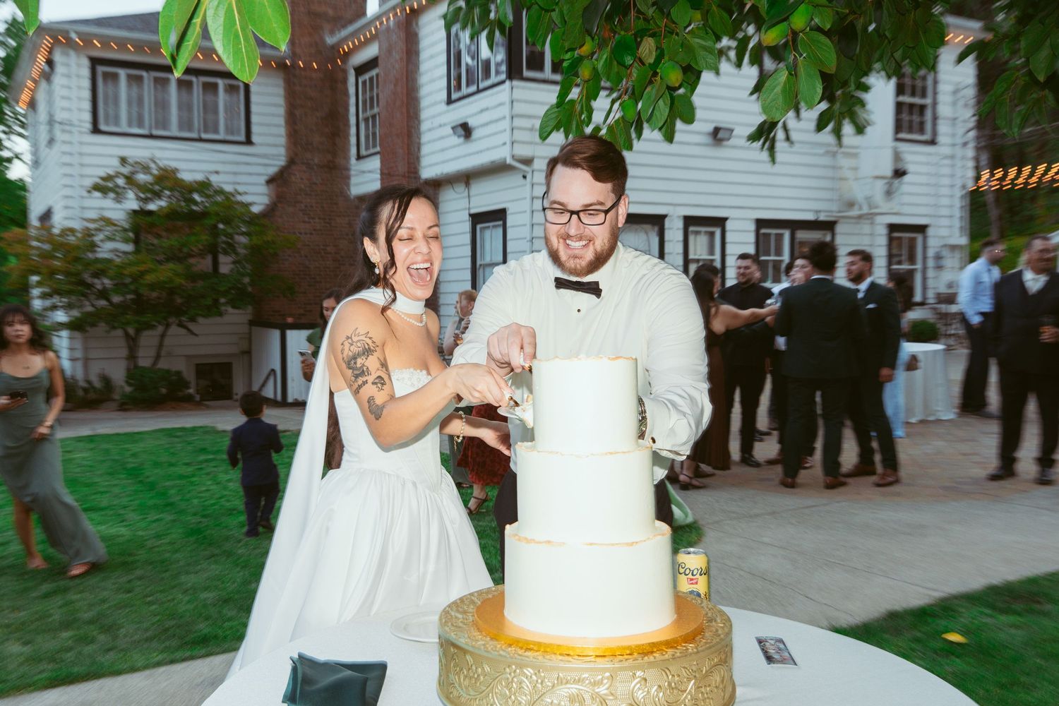 A couple cuts their elegant white wedding cake while guests watch in the background.