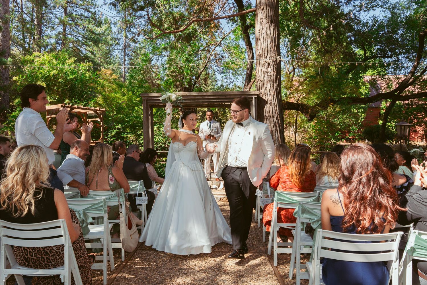 Outdoor wedding gathering with guests seated under wooden pergola surrounded by trees.