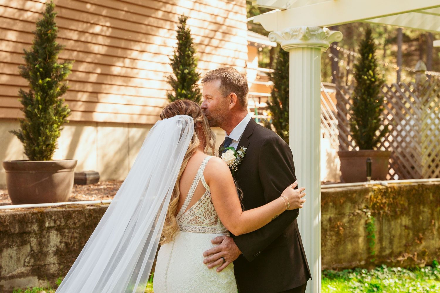 A couple shares an intimate first look moment under a white pergola surrounded by greenery.