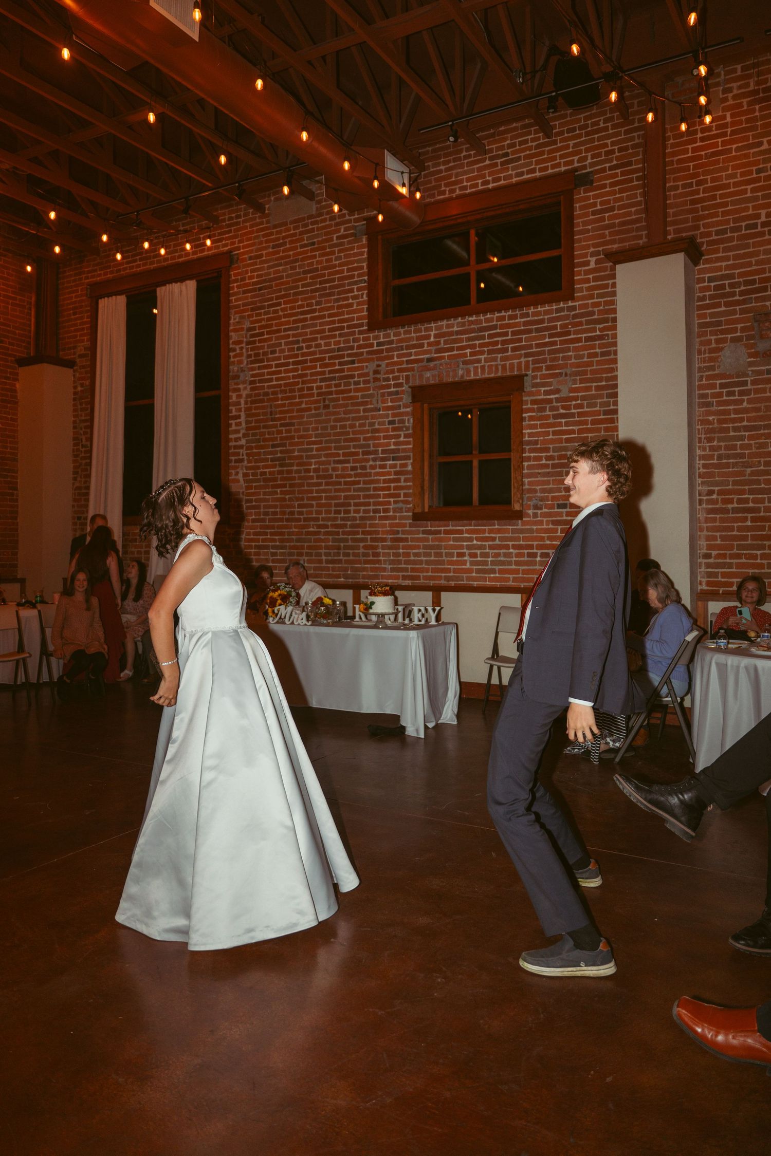 Wedding party dancing in a rustic brick venue with string lights overhead.