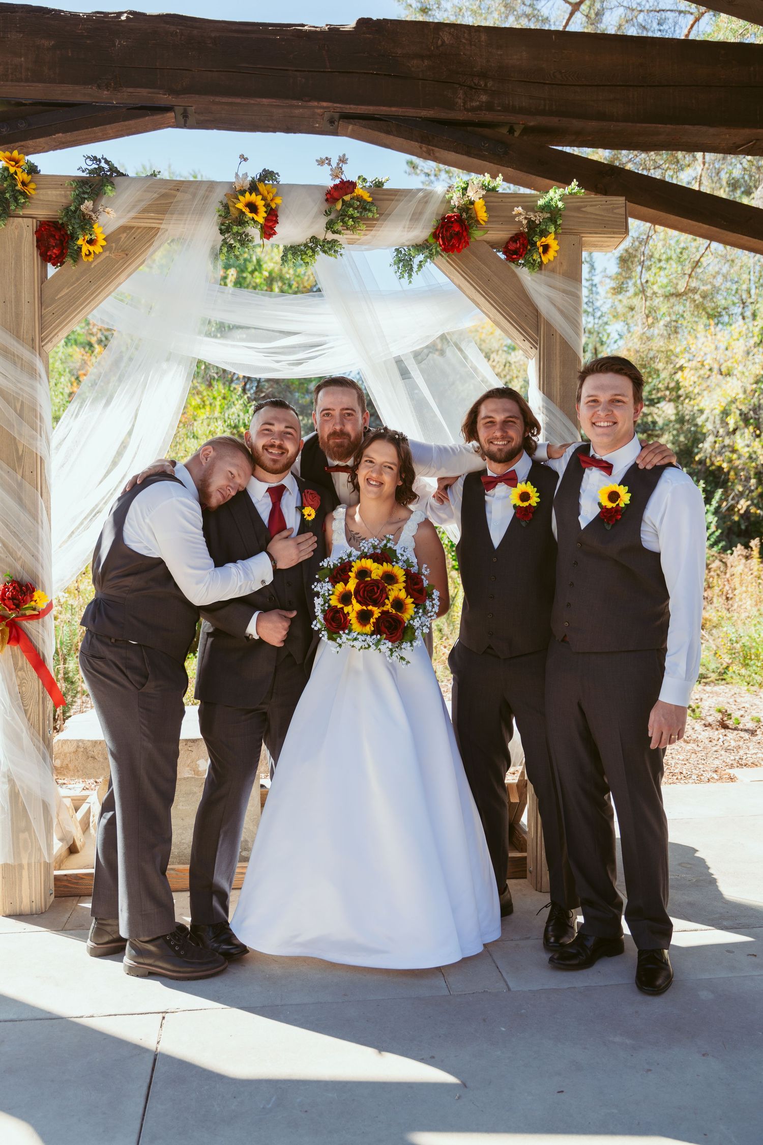 Wedding party in vests and formal attire poses with sunflower bouquets under outdoor pavilion.