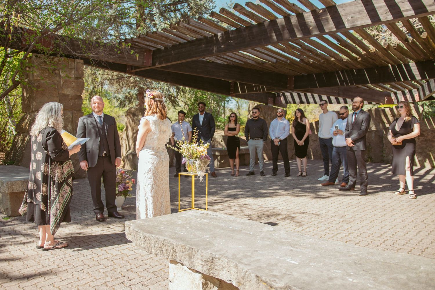 Wedding ceremony taking place under a rustic wooden pergola with guests seated in an outdoor garden setting.