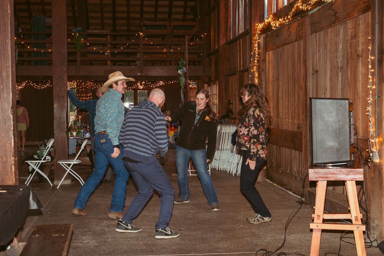 People dancing and celebrating in a wooden barn decorated with festive lights.