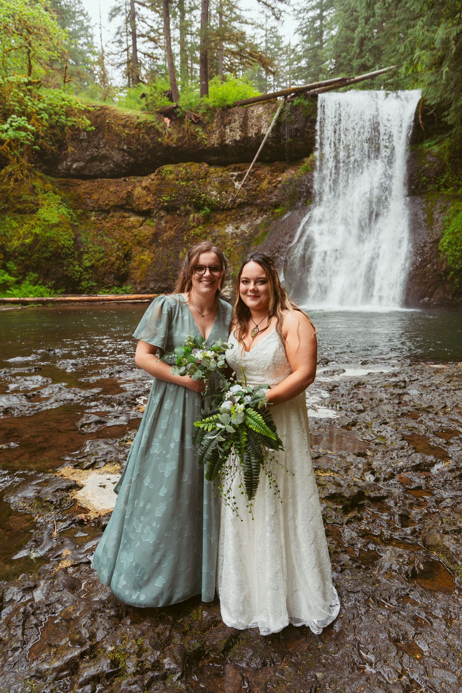 Wedding party in sage green and white dresses pose together near a cascading waterfall.