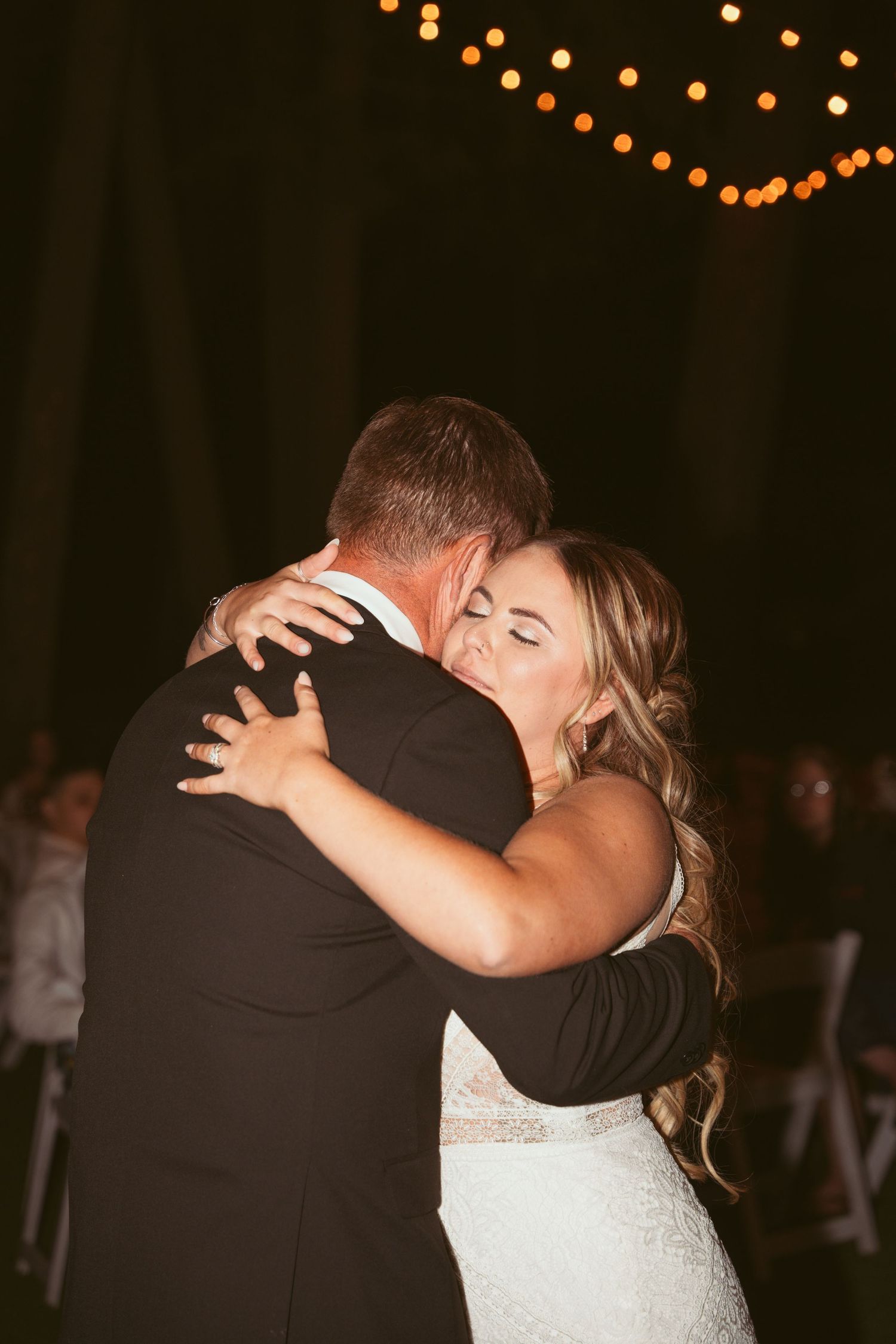 A tender embrace between newlyweds during their first dance, with string lights glowing softly overhead.