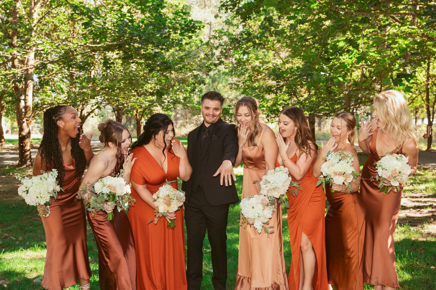 Large wedding party in orange dresses and dark suits poses together outdoors.