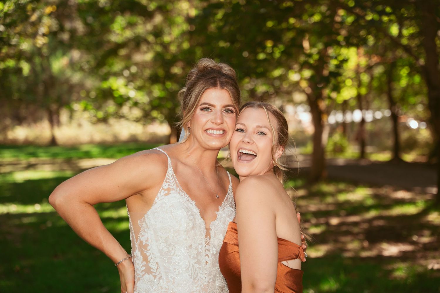 Wedding party in orange and white dresses pose together on a sunny lawn with trees.