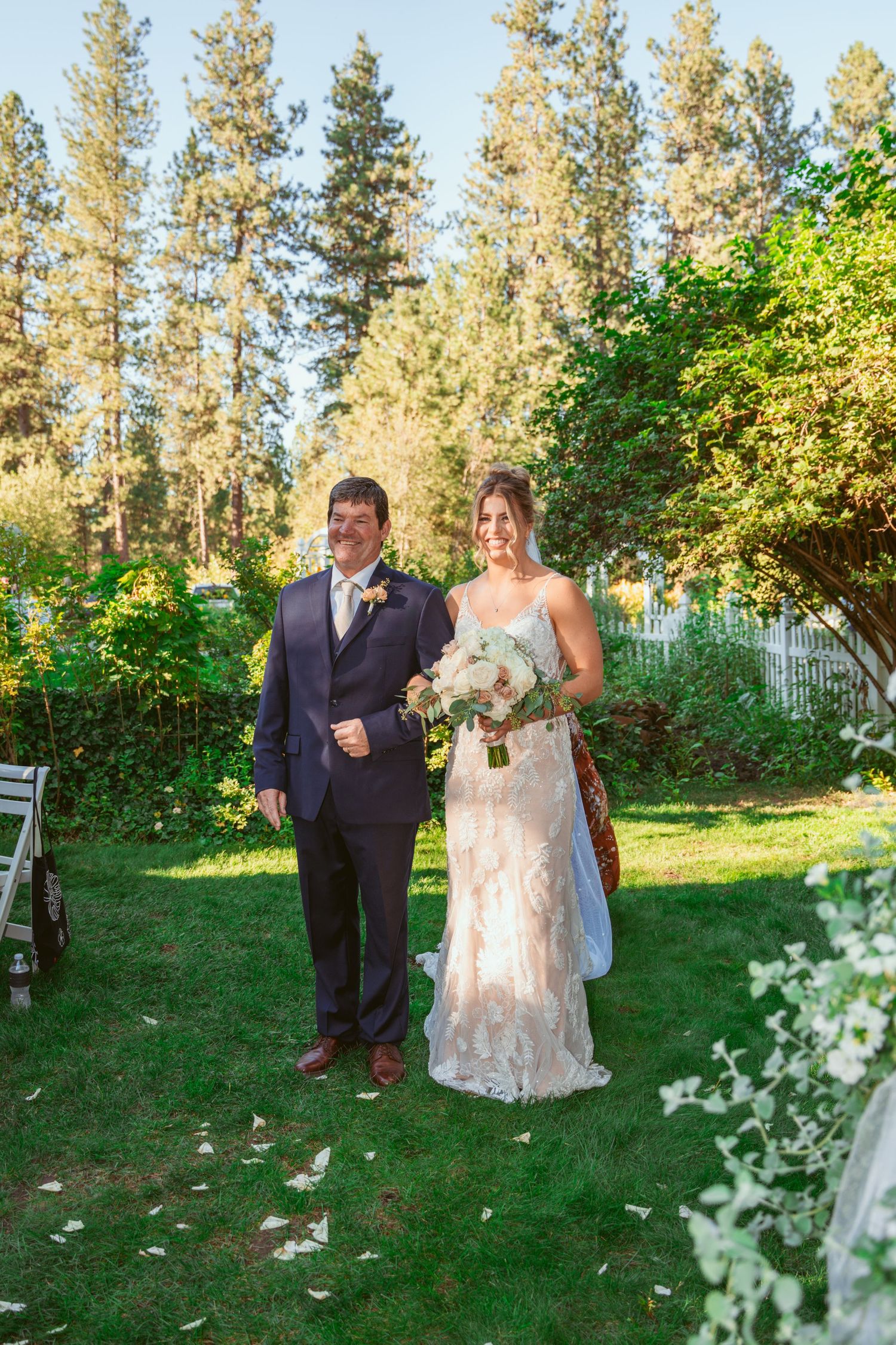 Wedding couple walks along a grassy path lined with tall pine trees during an outdoor ceremony.