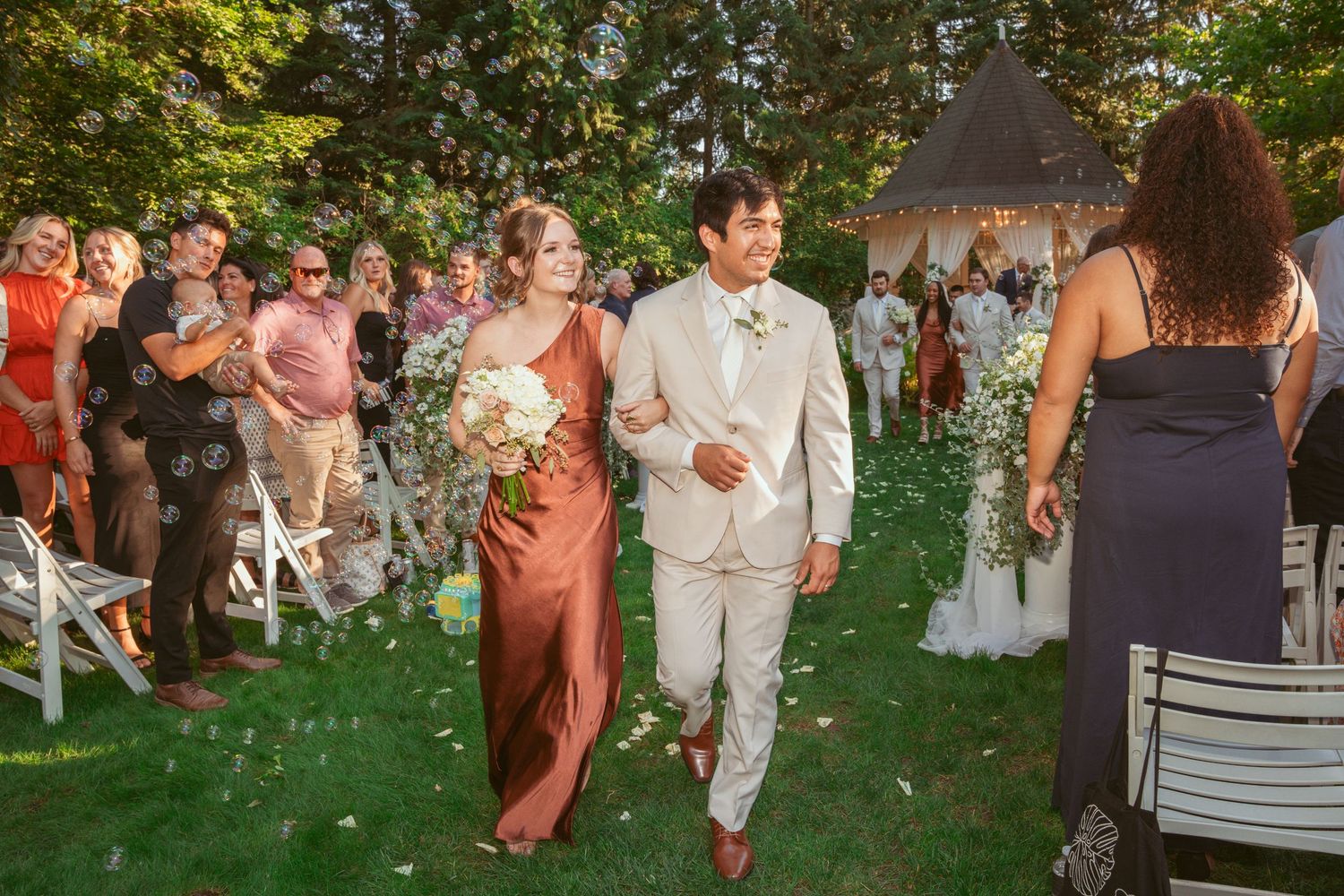 Wedding guests in formal attire gathering on a lush green lawn during sunset.