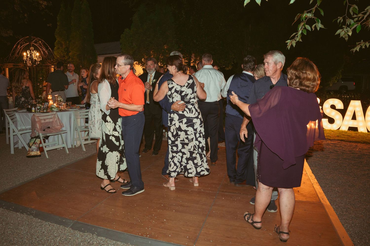 Evening outdoor wedding reception area with guests mingling near decorative lighting.