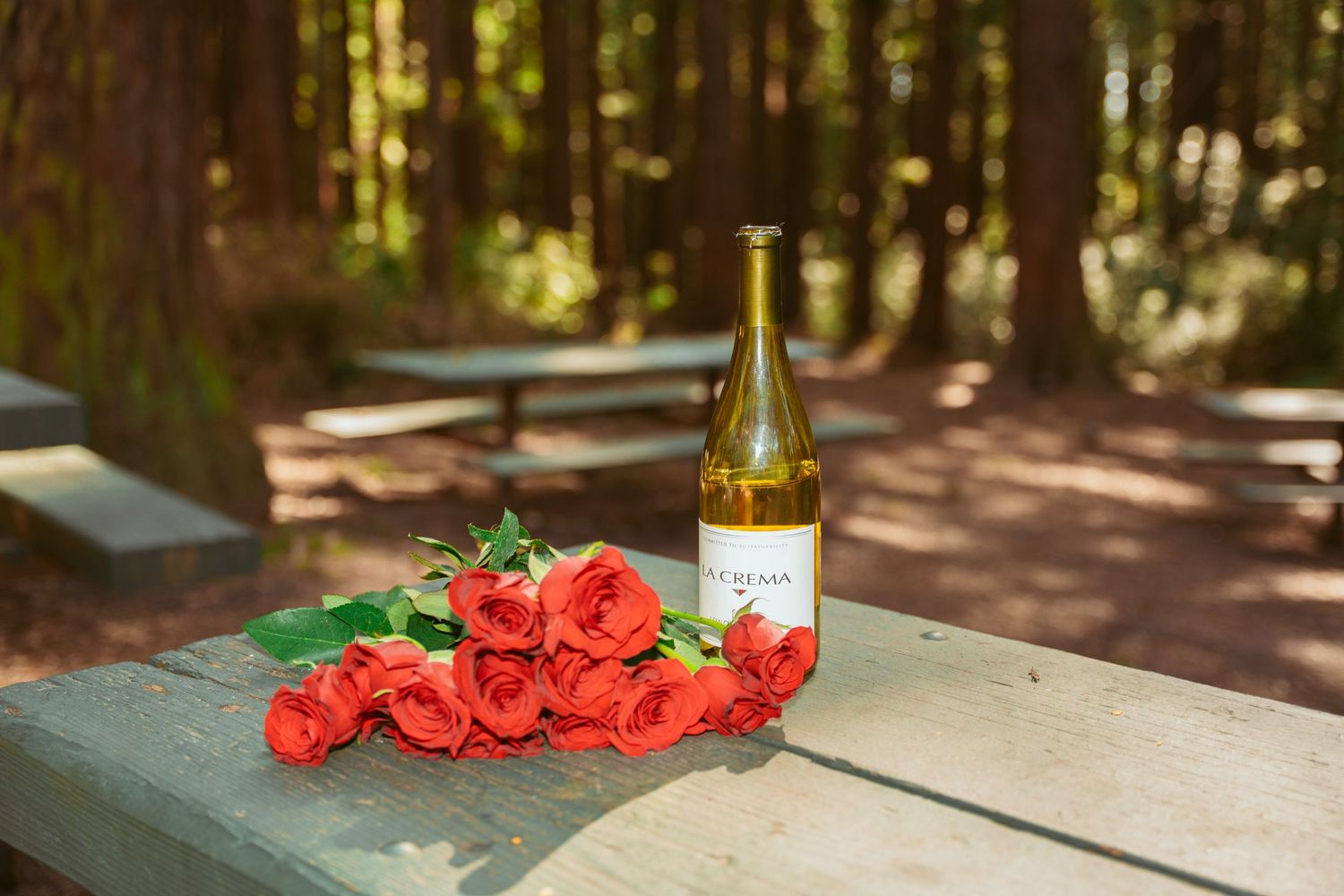 Red roses and wine bottles arranged on an outdoor table in a forest setting.