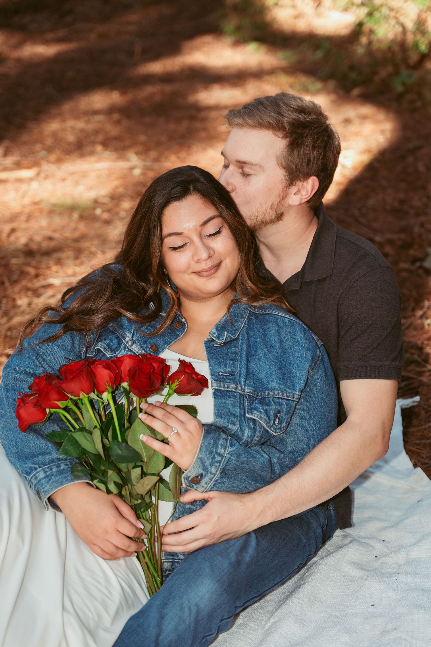 Romantic couple sharing intimate moments on a blanket with red roses in a forest setting.