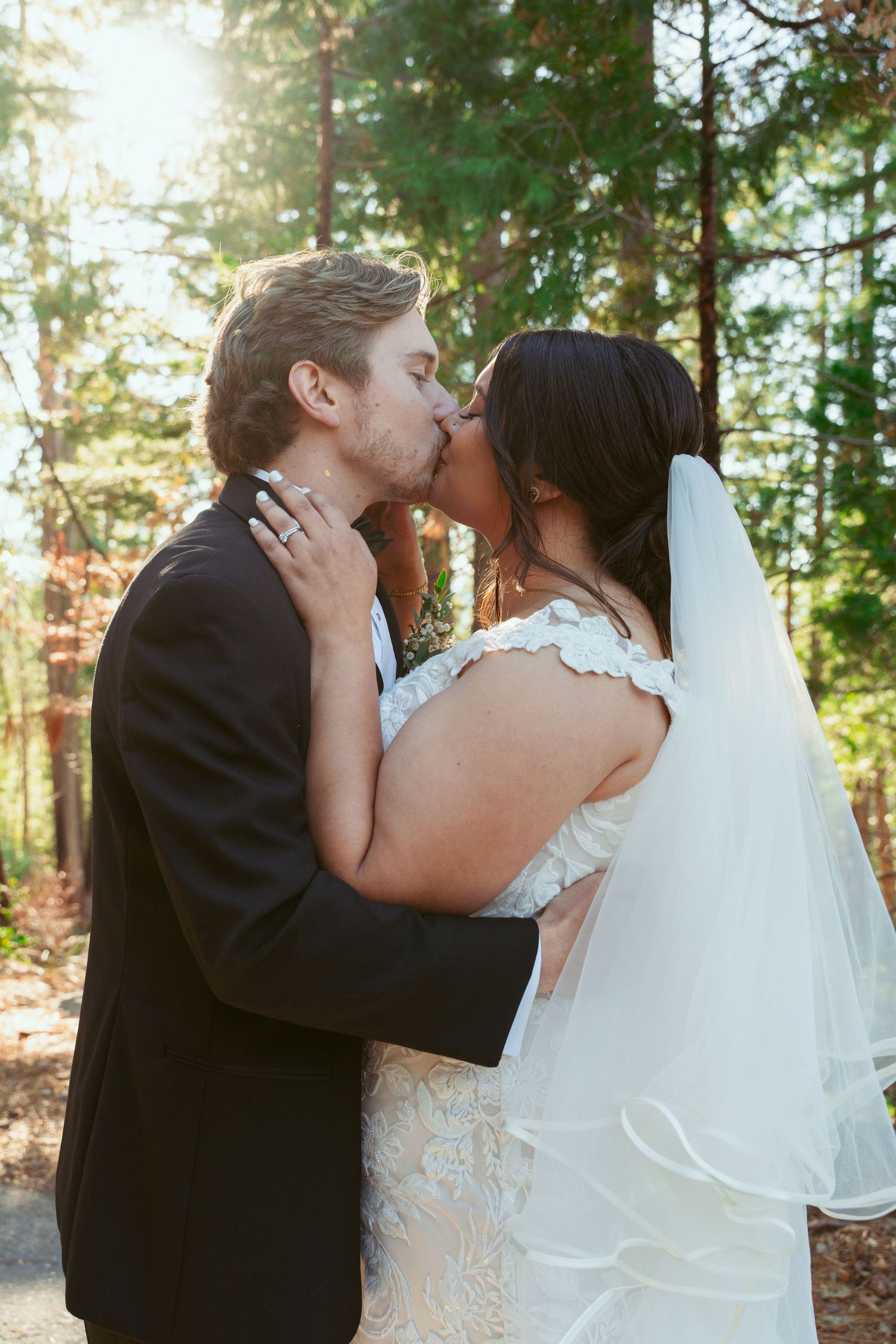 Intimate wedding portrait with sunlight filtering through pine trees in the background.