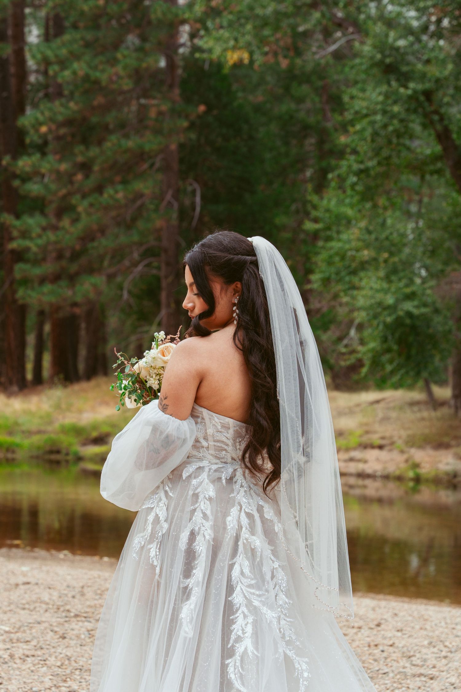 Elegant lace wedding dress with flowing sleeves and veil captured in a scenic forest setting near a lake.