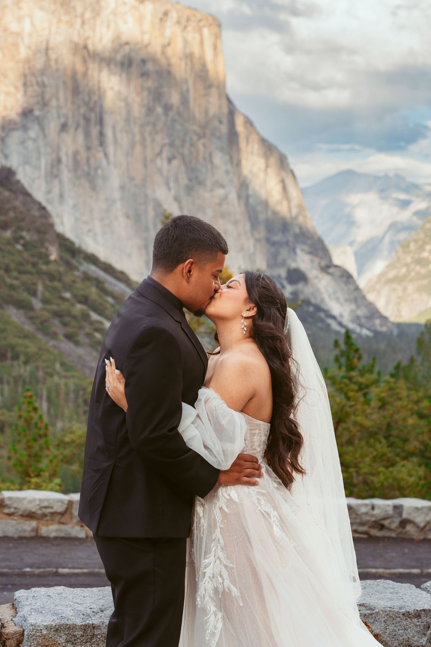 Newlyweds embracing against stunning Yosemite Valley backdrop with El Capitan visible in distance.
