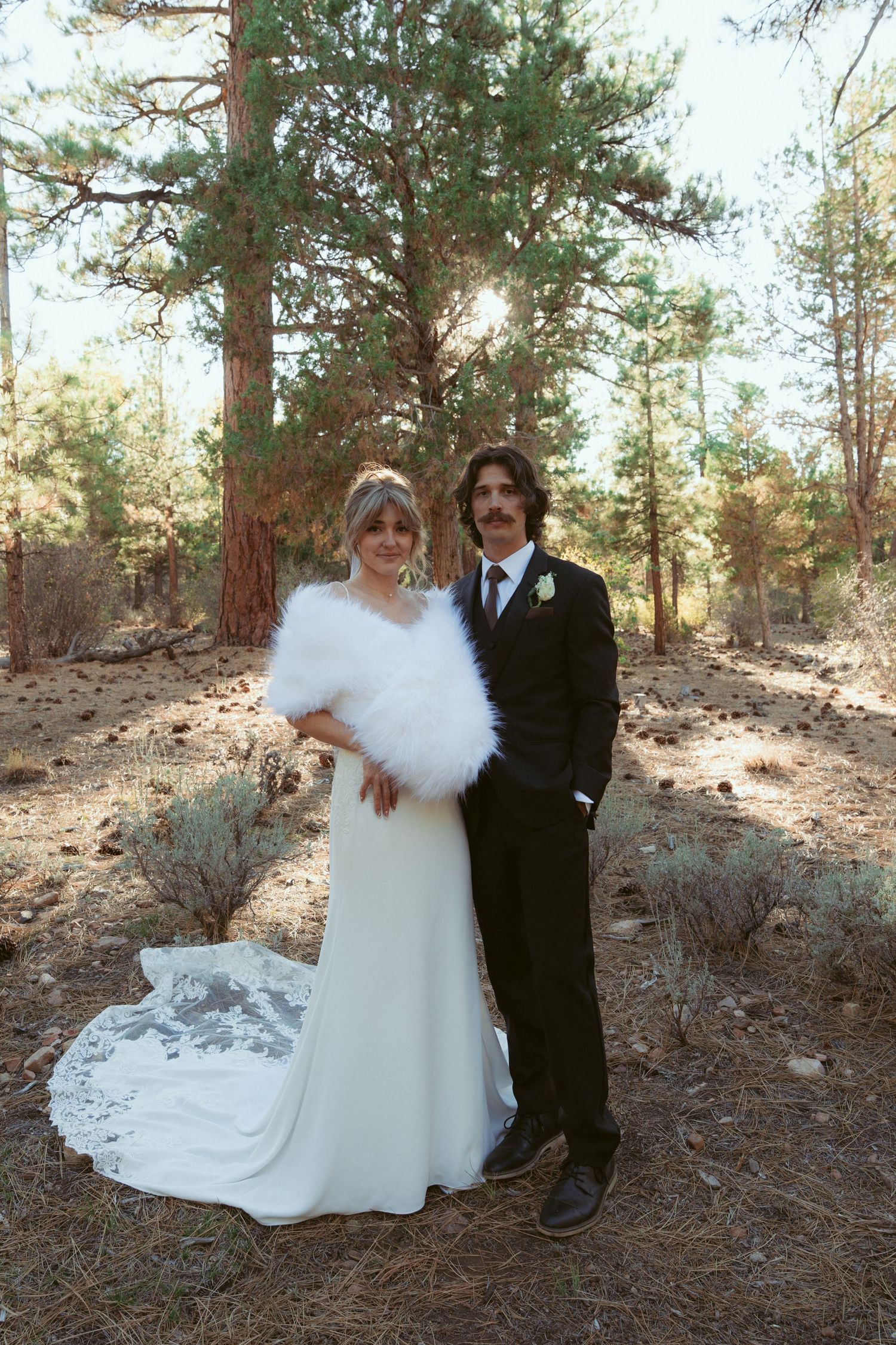 A bride in a white dress and fur shawl poses with her partner in a pine forest.