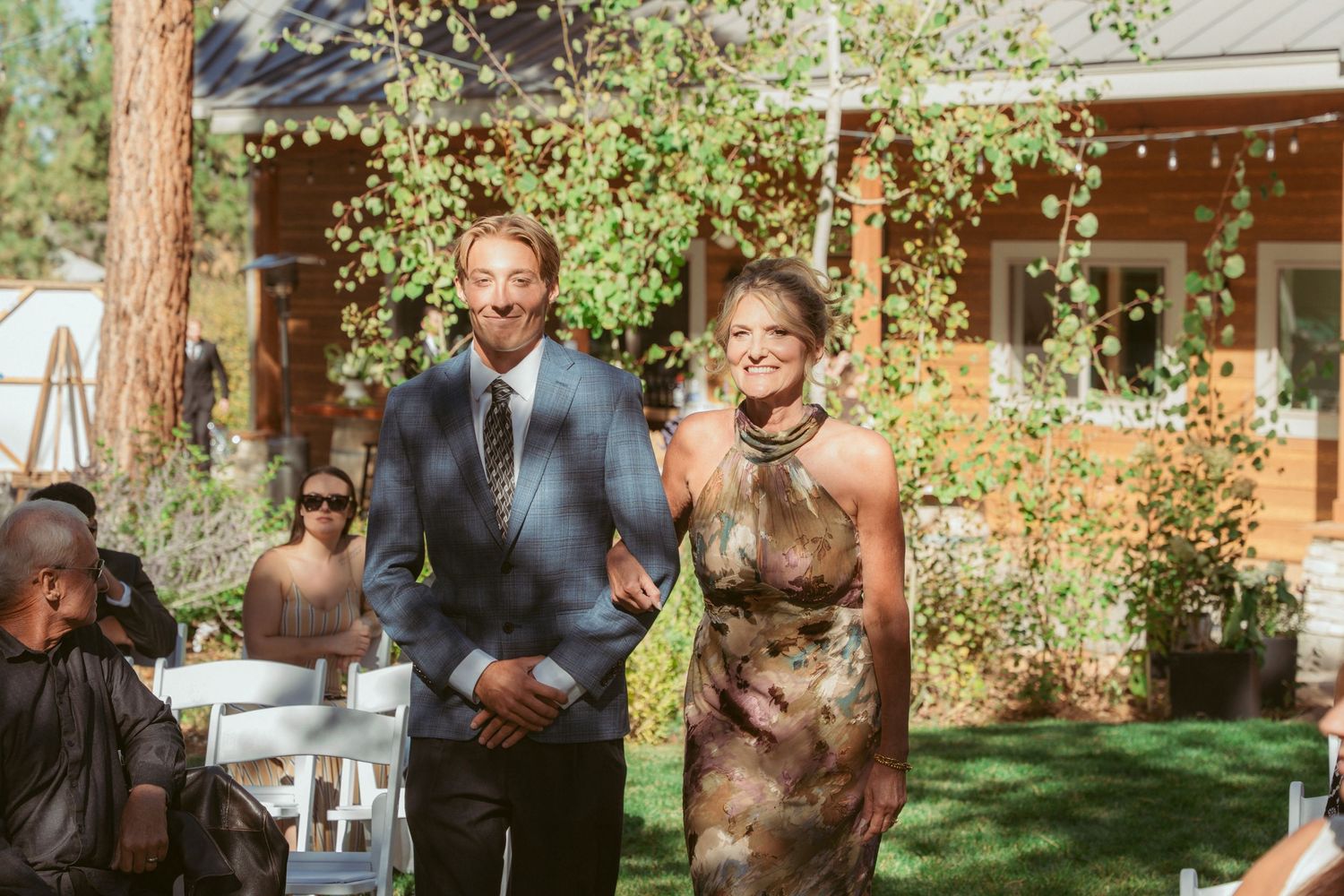 Wedding party members walk down an outdoor aisle with a rustic cabin in the background.