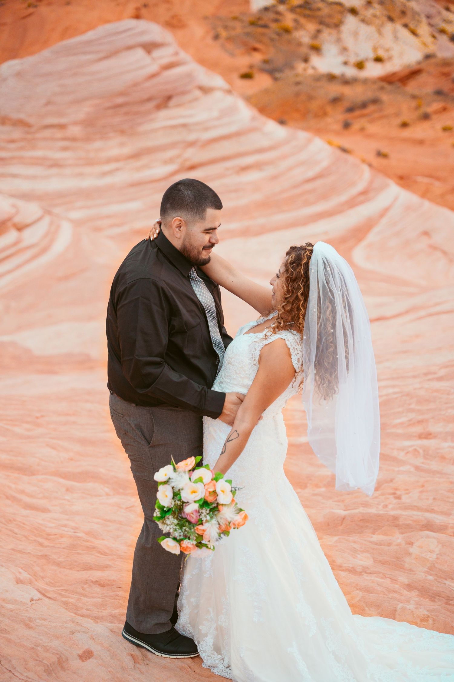 Bride in white lace dress holds bouquet while posing on red sandstone formations.