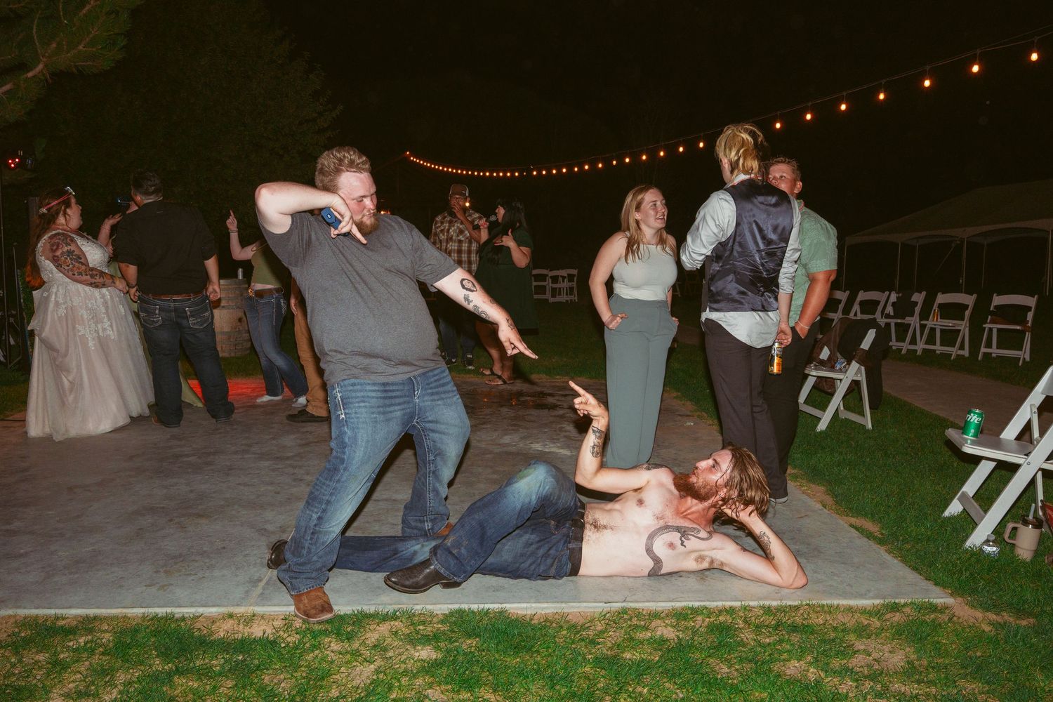Wedding guests perform a fun dance move in a line at an outdoor reception under string lights.