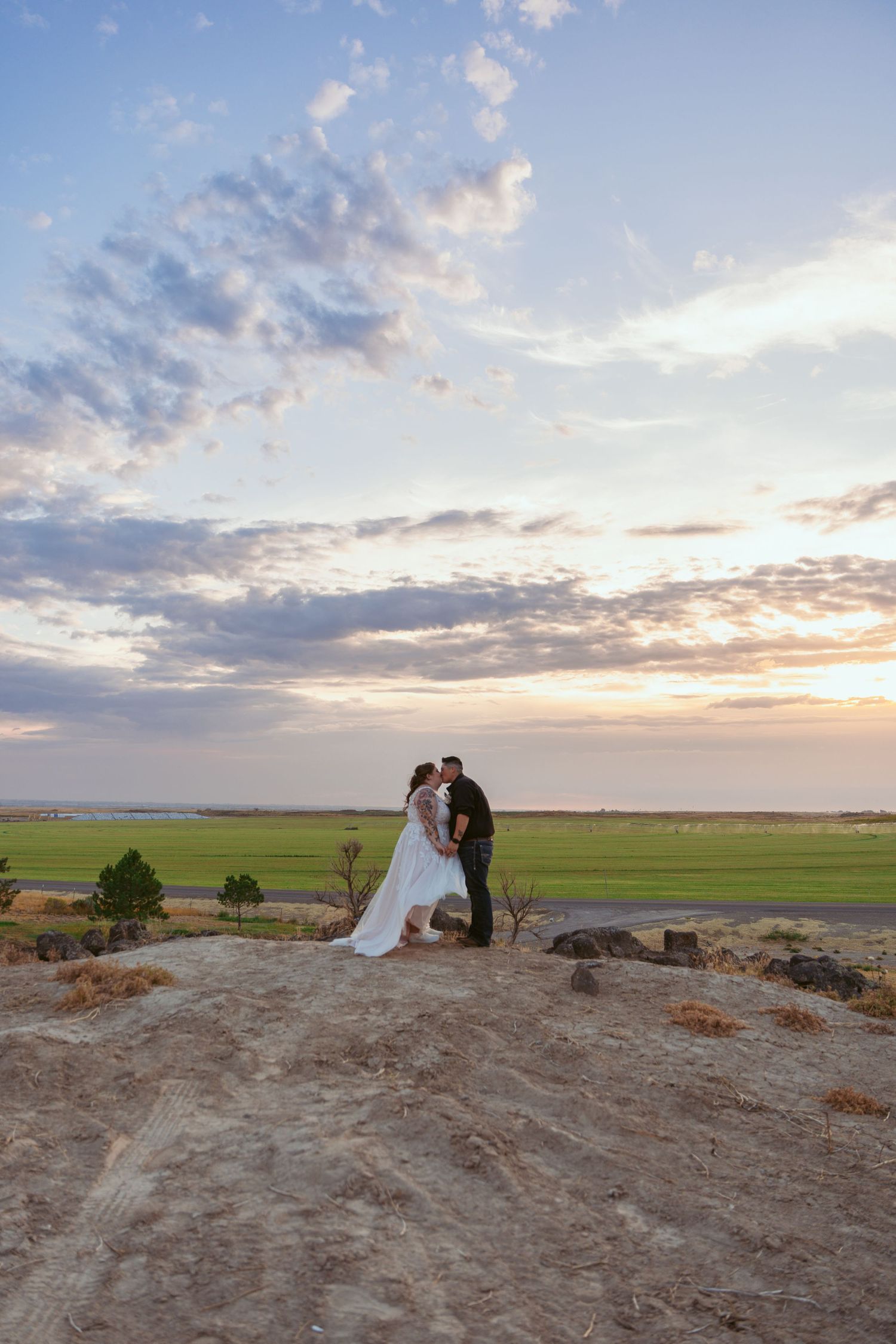 Romantic sunset wedding photo on a hilltop overlooking vast green fields and dramatic sky.