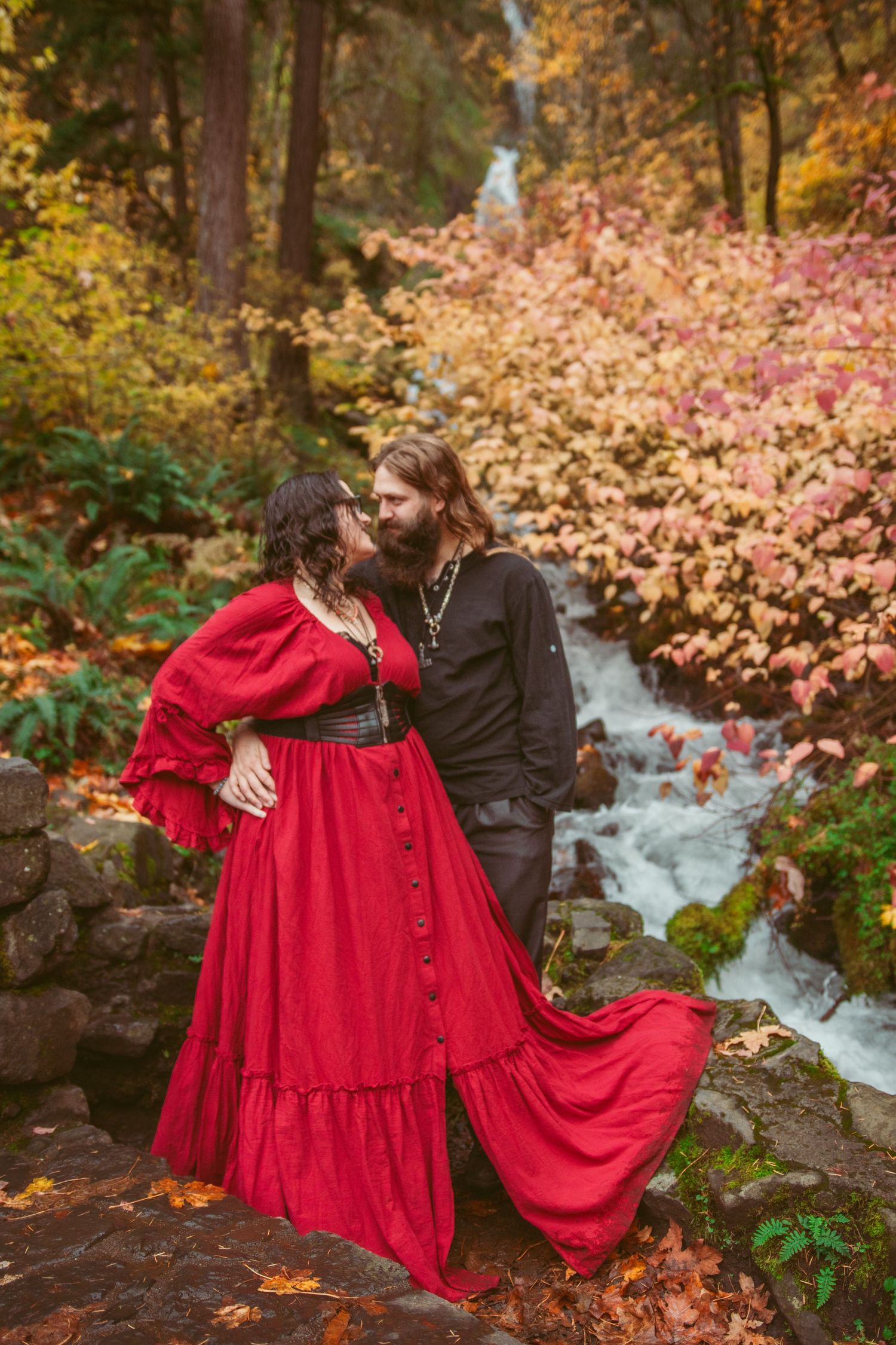 A couple embraces by a waterfall surrounded by autumn foliage, with one wearing a flowing red dress.