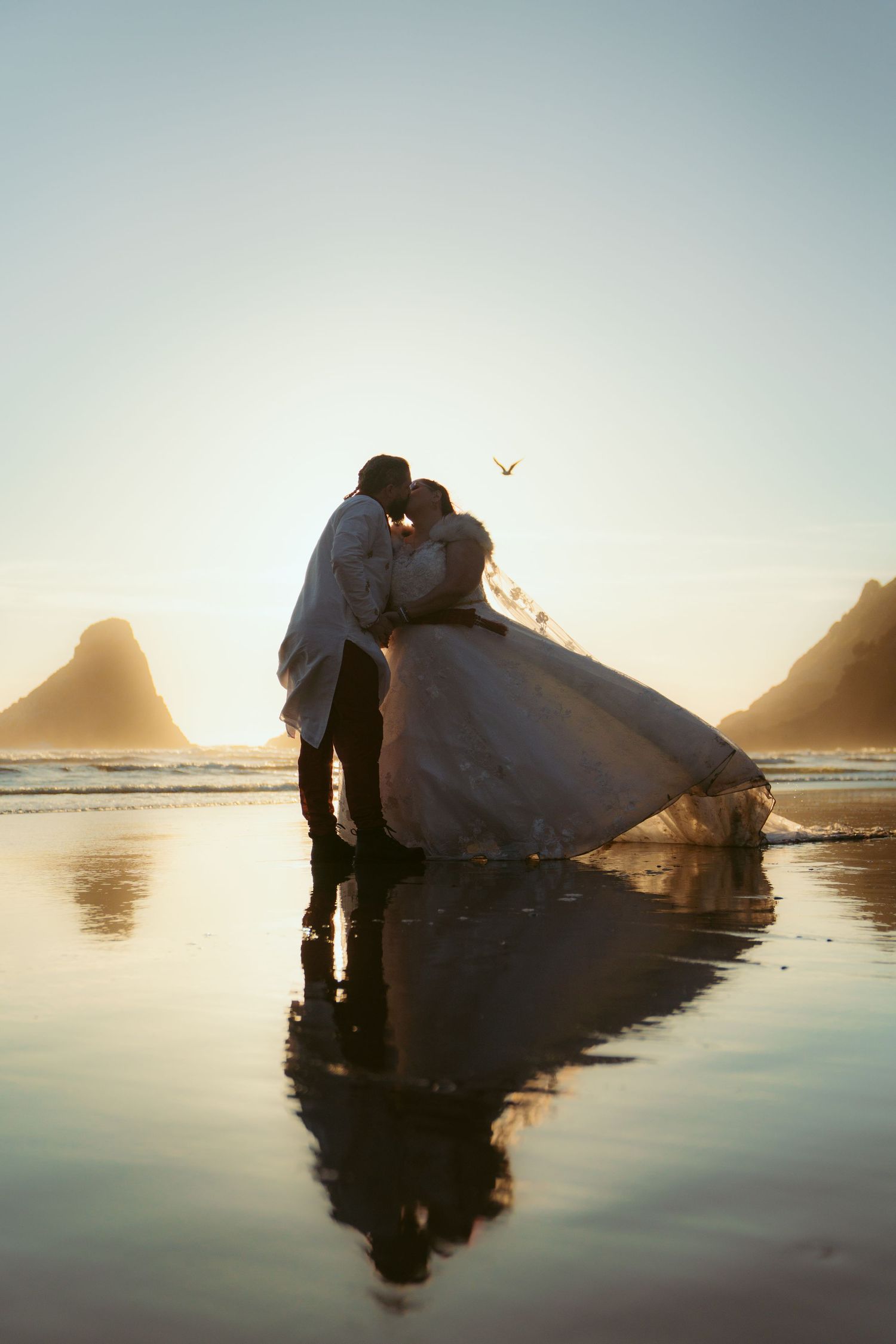 Couple embracing on reflective beach at golden hour with dramatic coastal landscape.