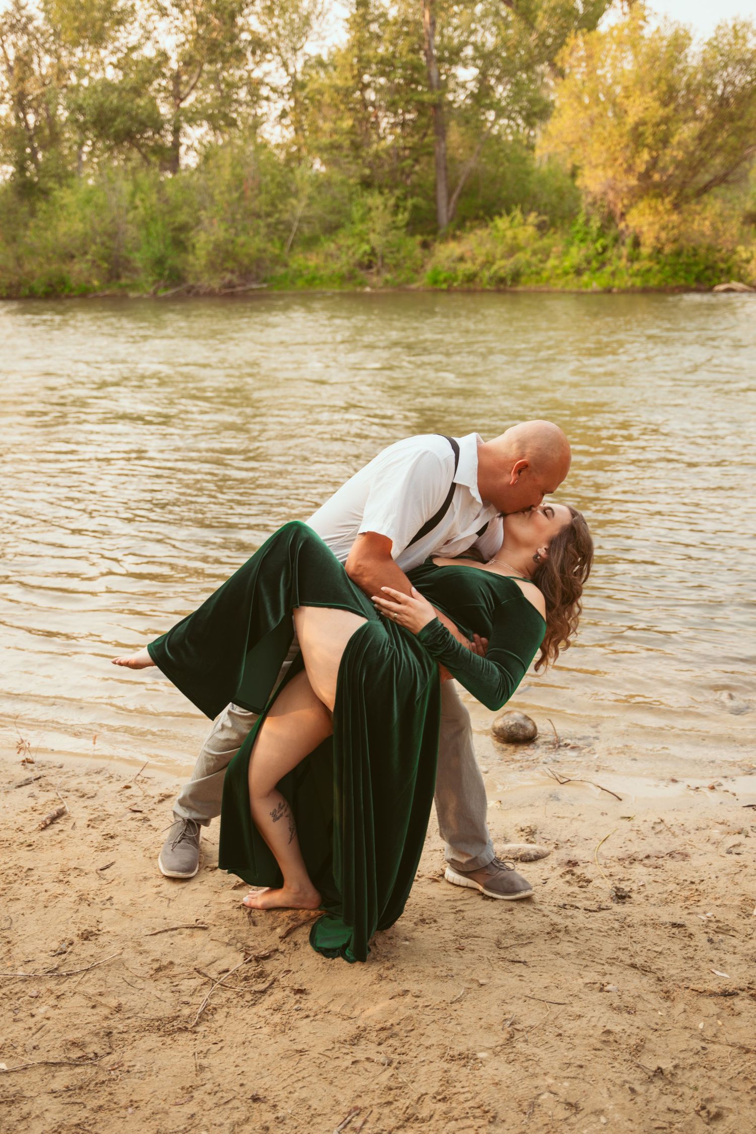 Dancing couple performs a dramatic dip pose on a sandy riverbank surrounded by fall trees.
