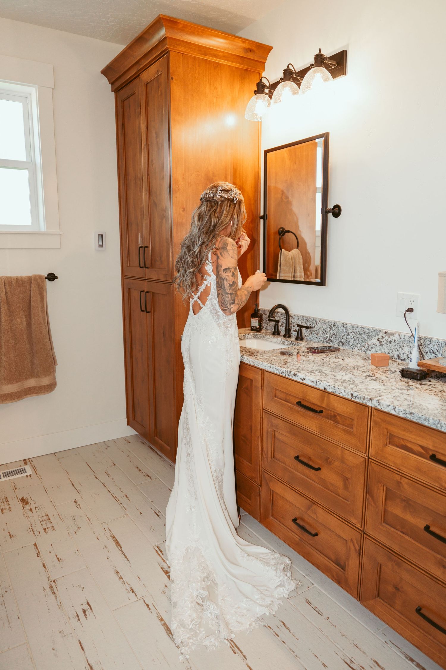 Bride in flowing white dress standing in bathroom with wooden cabinetry.
