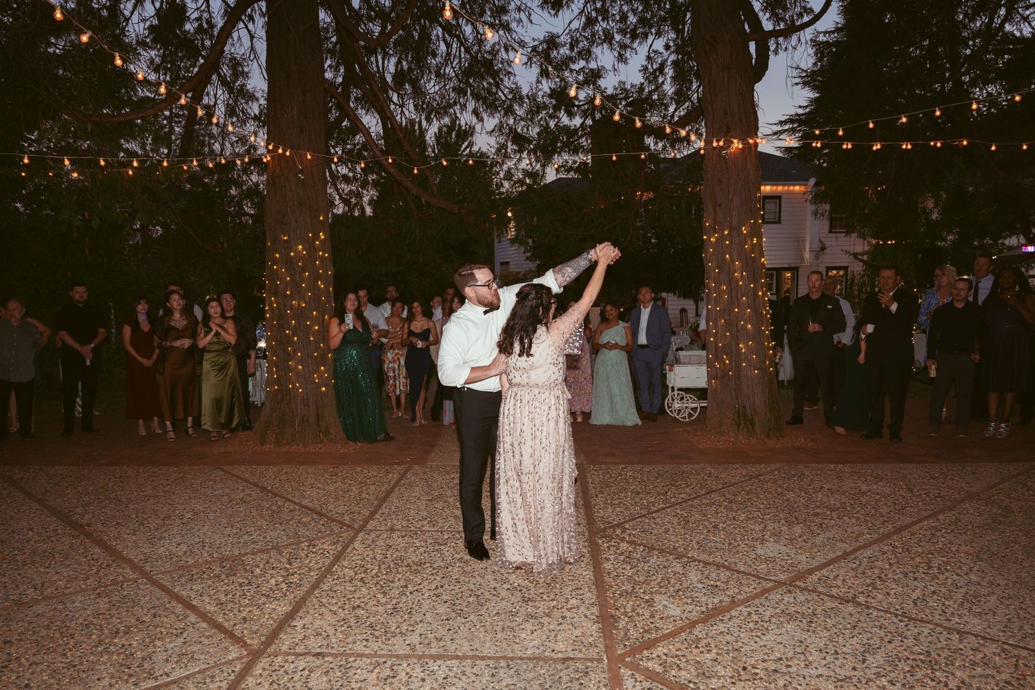 String lights illuminate an outdoor wedding dance floor surrounded by tall trees at night.