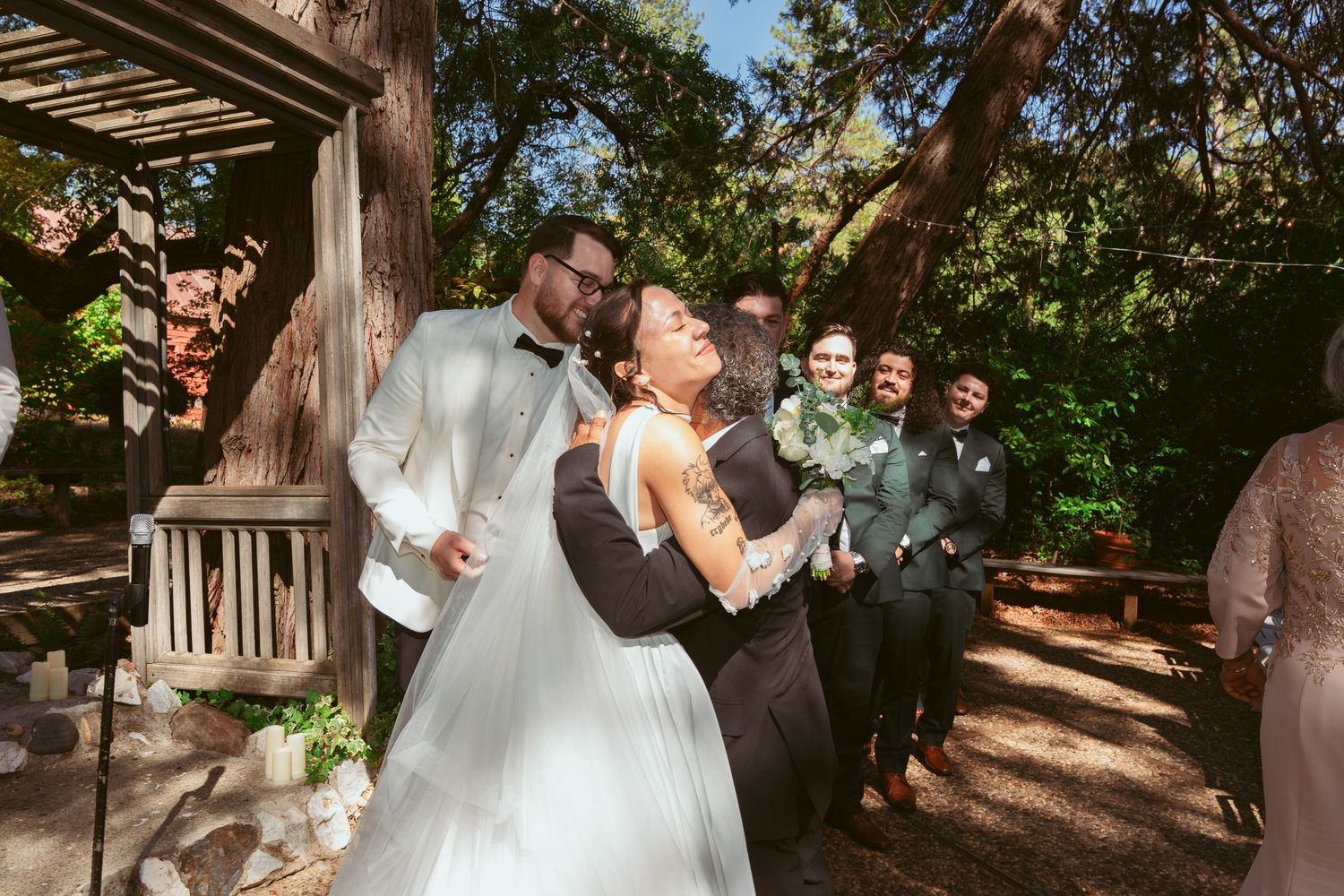 Wedding celebration moment captured under wooden pergola with natural forest backdrop.