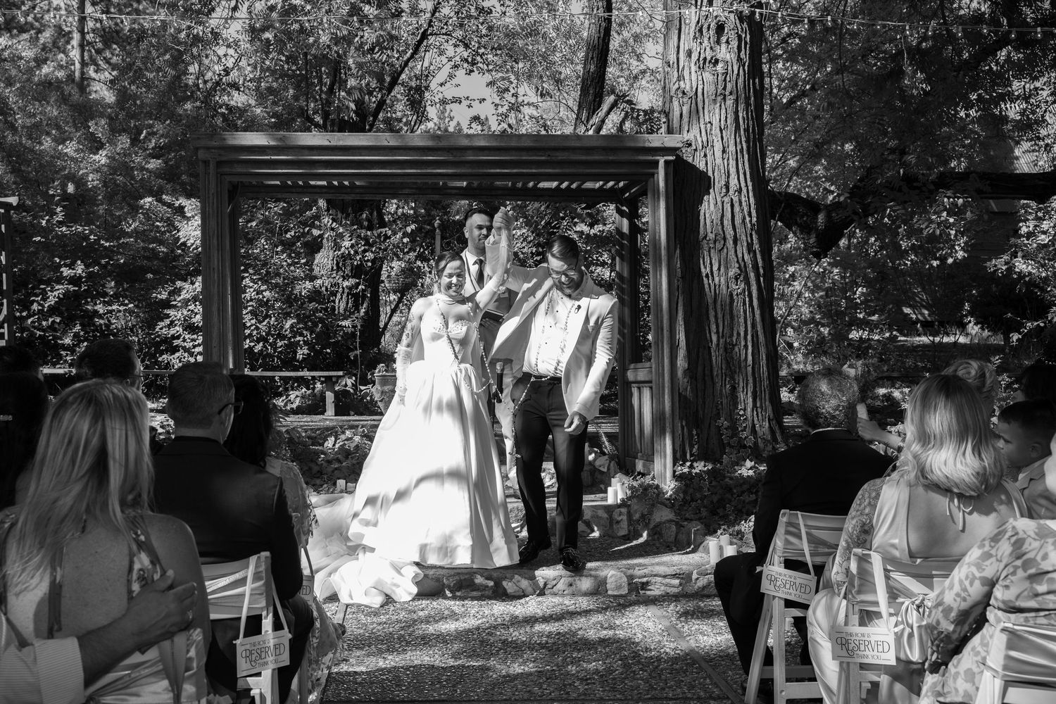 A black and white photograph of an outdoor wedding ceremony in a wooded setting with a rustic wooden arch.