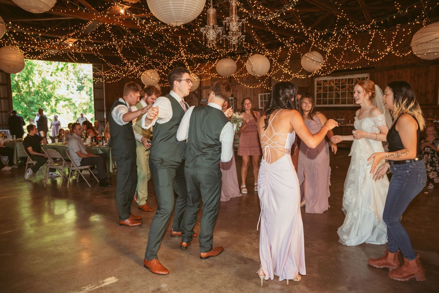 Wedding guests perform a line dance in a rustic barn decorated with paper lanterns and fairy lights.