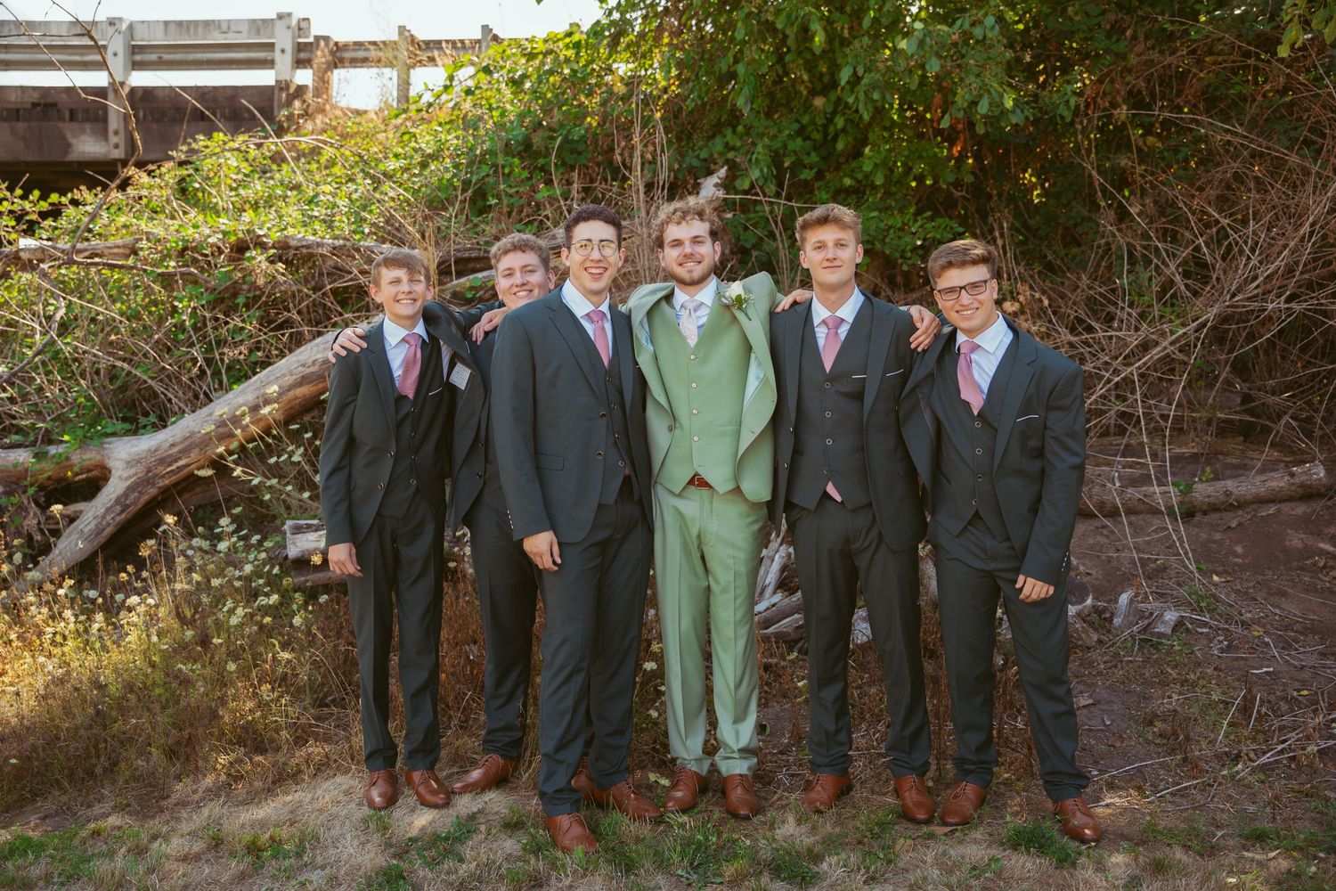 Groomsmen in dark suits stand together for formal photo against natural outdoor setting.