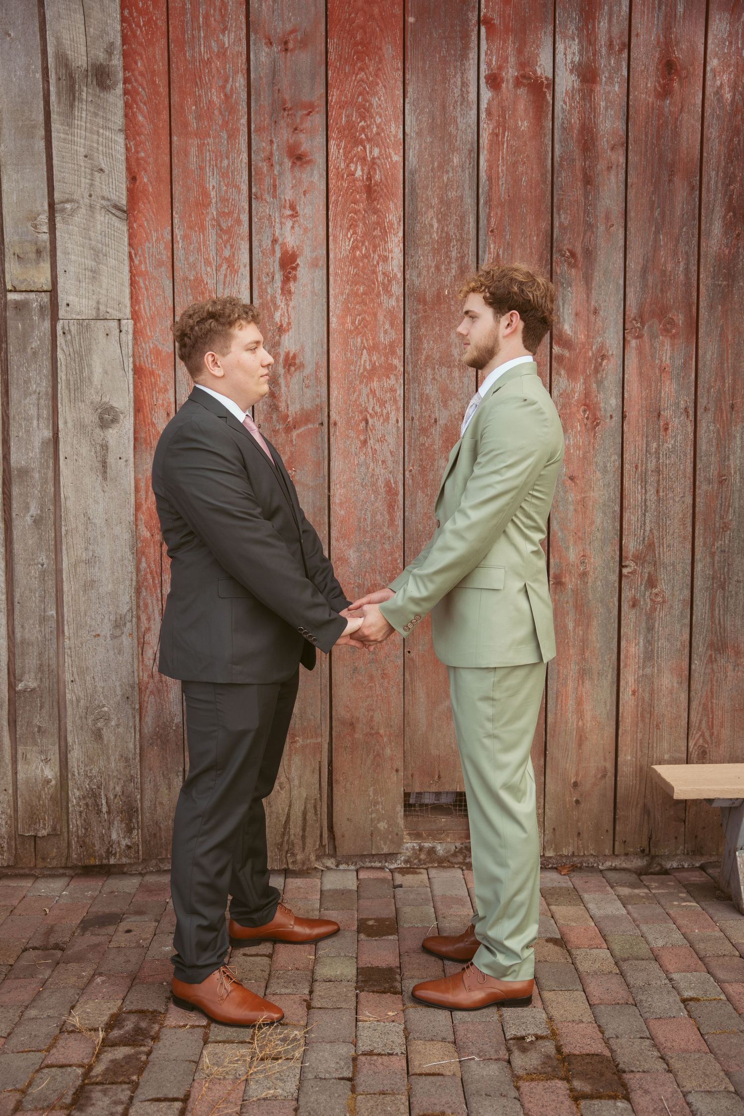 Two men in contrasting suits playfully pose in a mock fight sequence against a rustic barn wall backdrop.