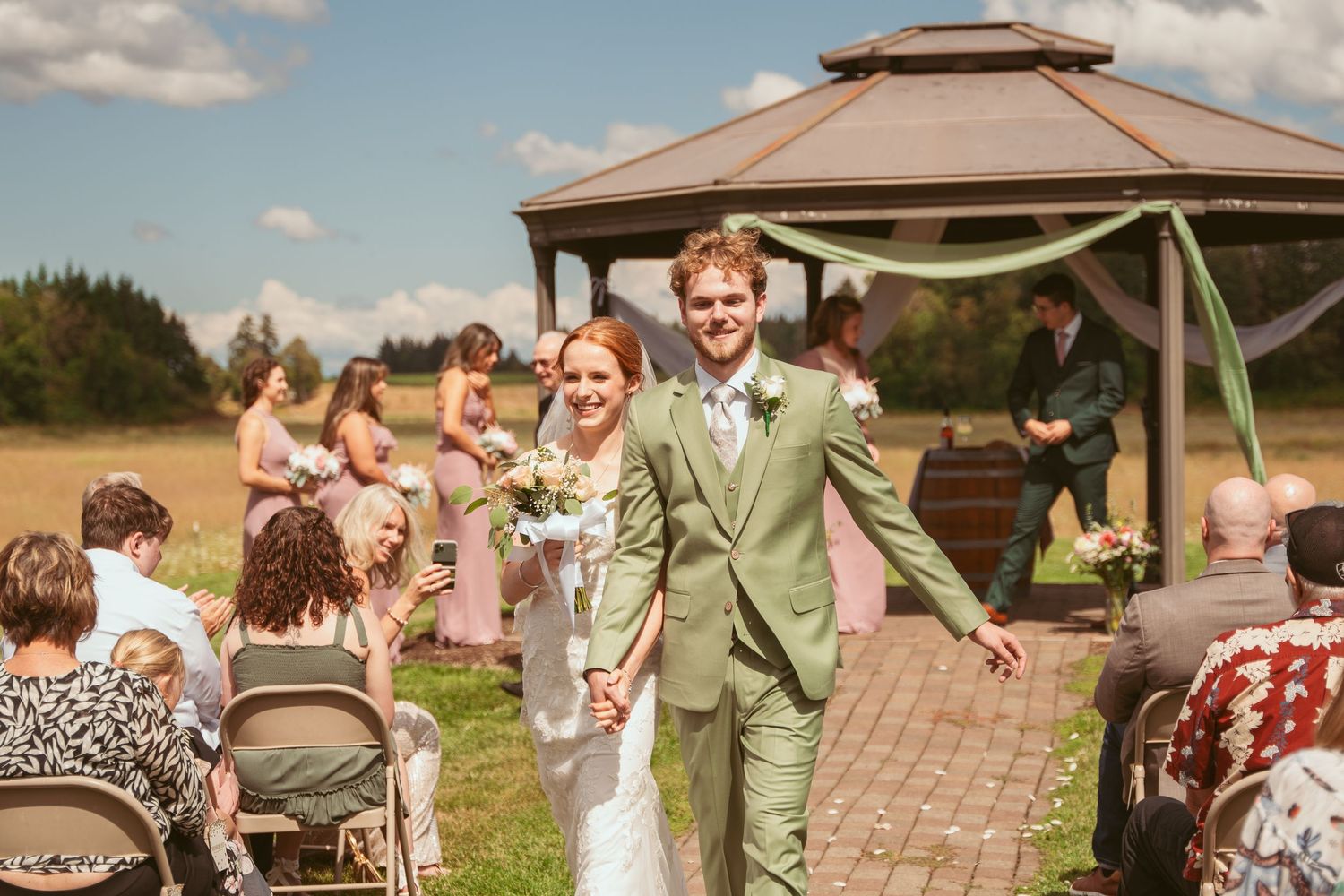 Joyful couple exits down brick aisle after outdoor gazebo ceremony with seated guests.