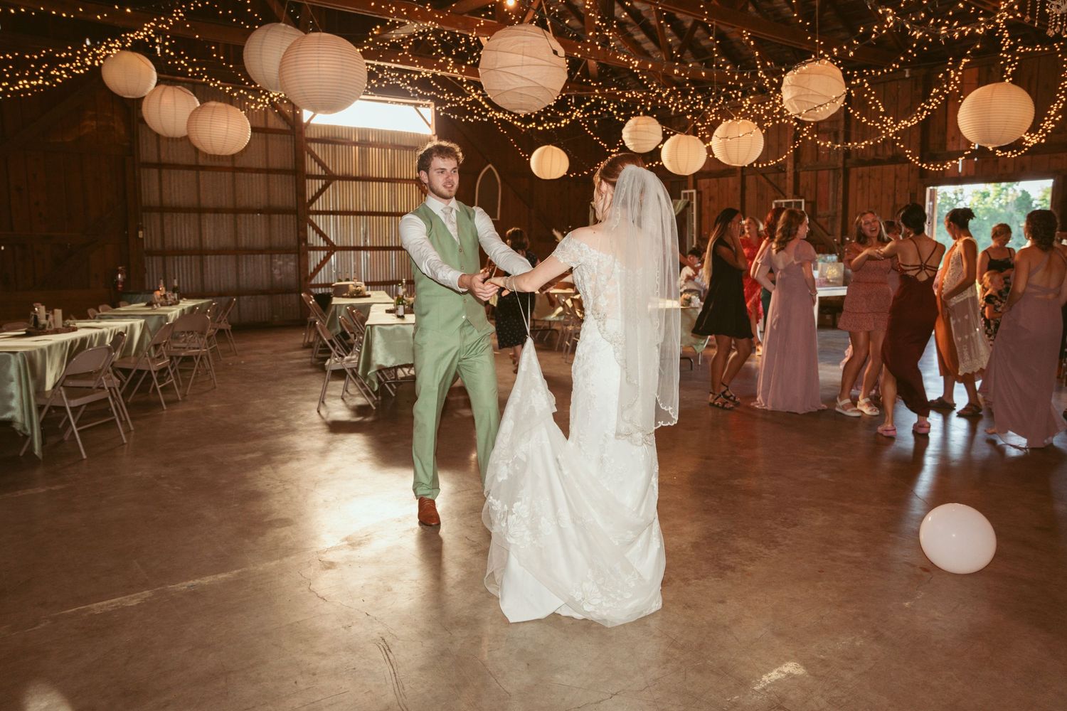Couple dancing with wedding guests in a barn venue decorated with string lights and lanterns.