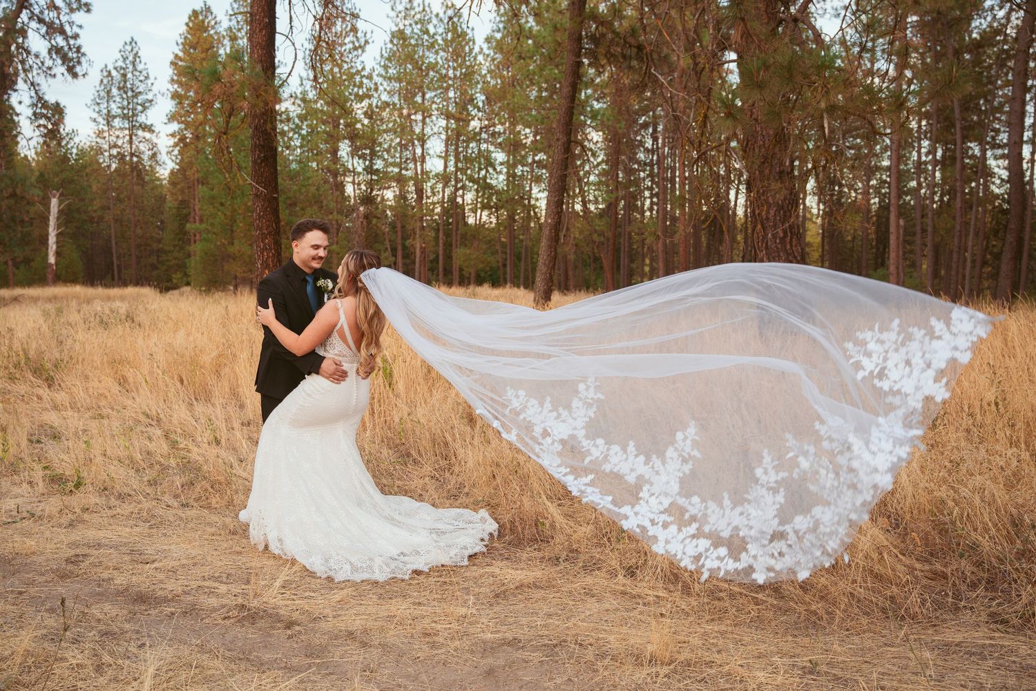 A bride in a flowing white wedding dress poses in a rustic outdoor setting with tall pine trees and golden grass.