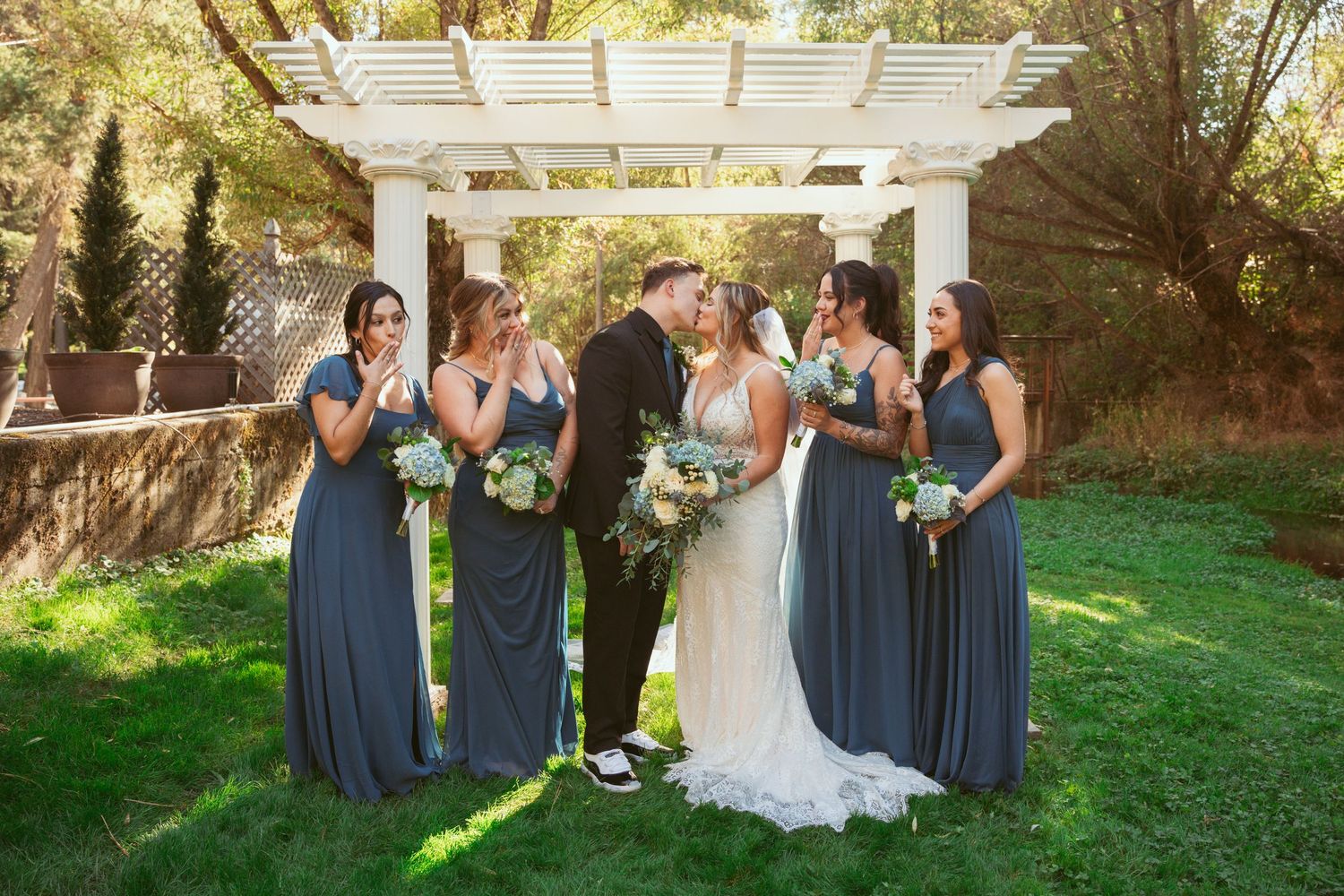 Wedding party in navy blue dresses gather under a white pergola for formal photos.