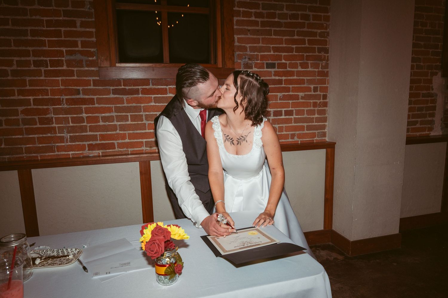Wedding couple at signing table with colorful flower bouquet in brick venue.