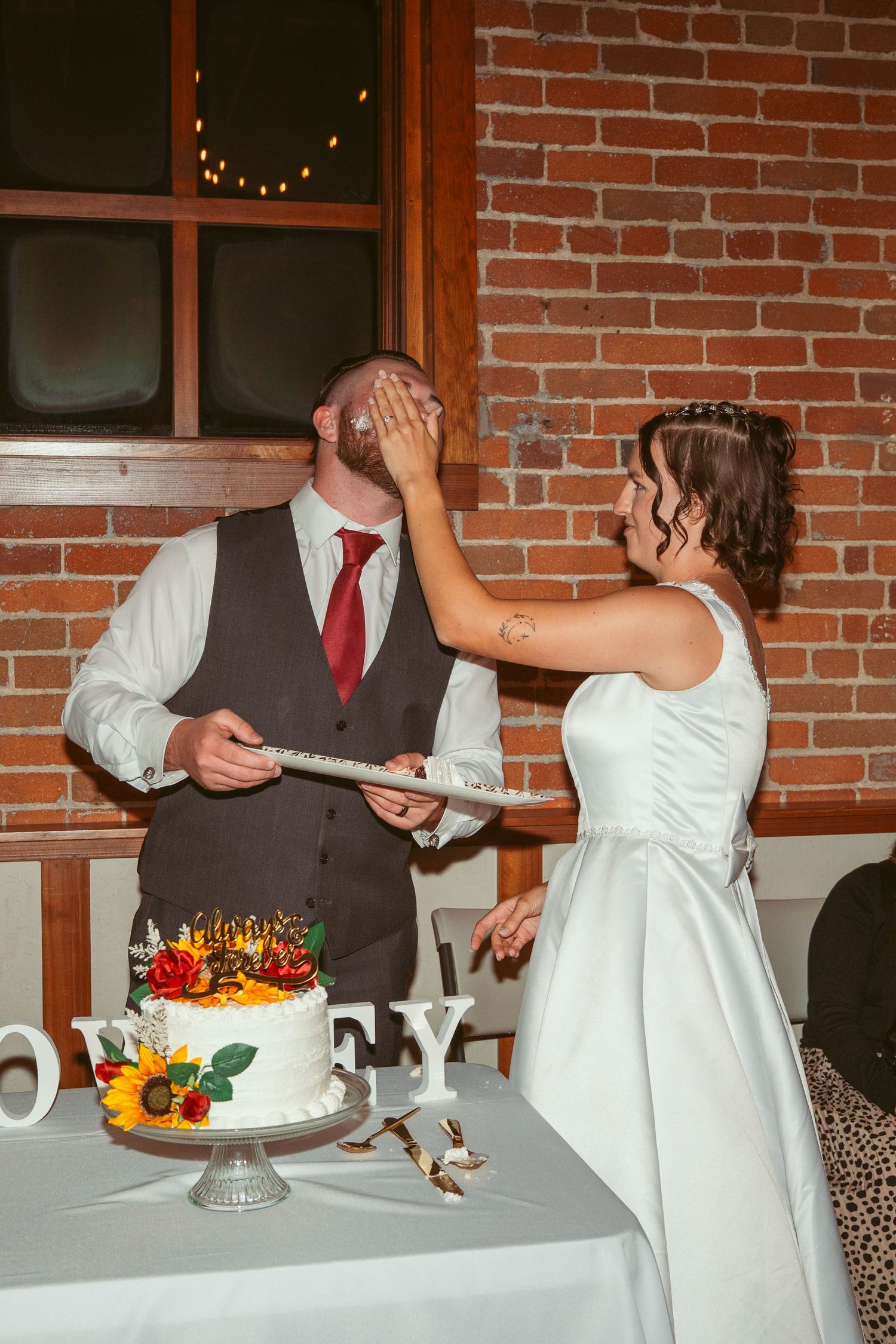 A couple at their wedding reception playfully feed each other cake in front of a brick wall backdrop.