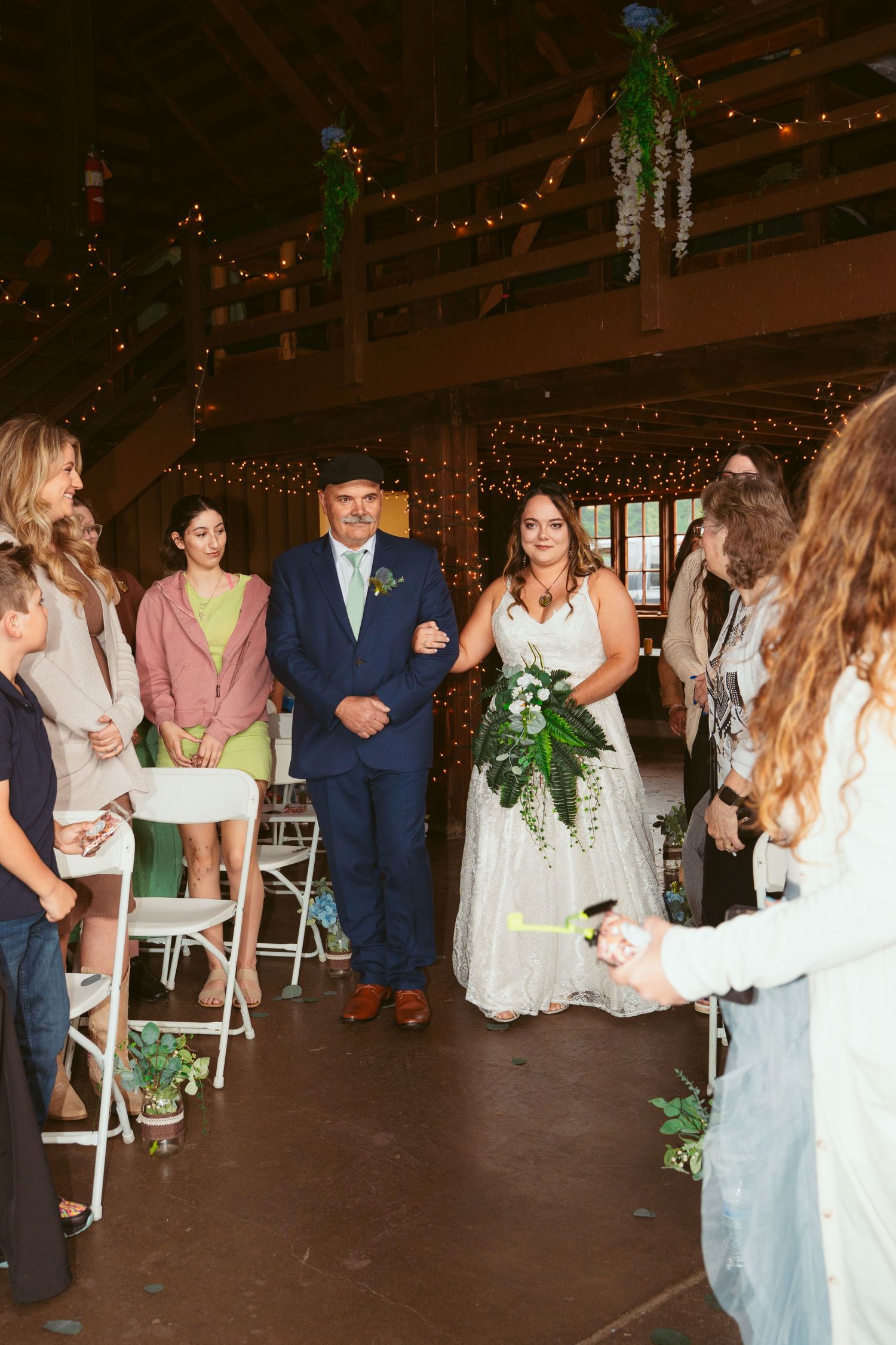 Wedding party in navy suits and light blue dresses gather inside rustic barn venue with string lights.