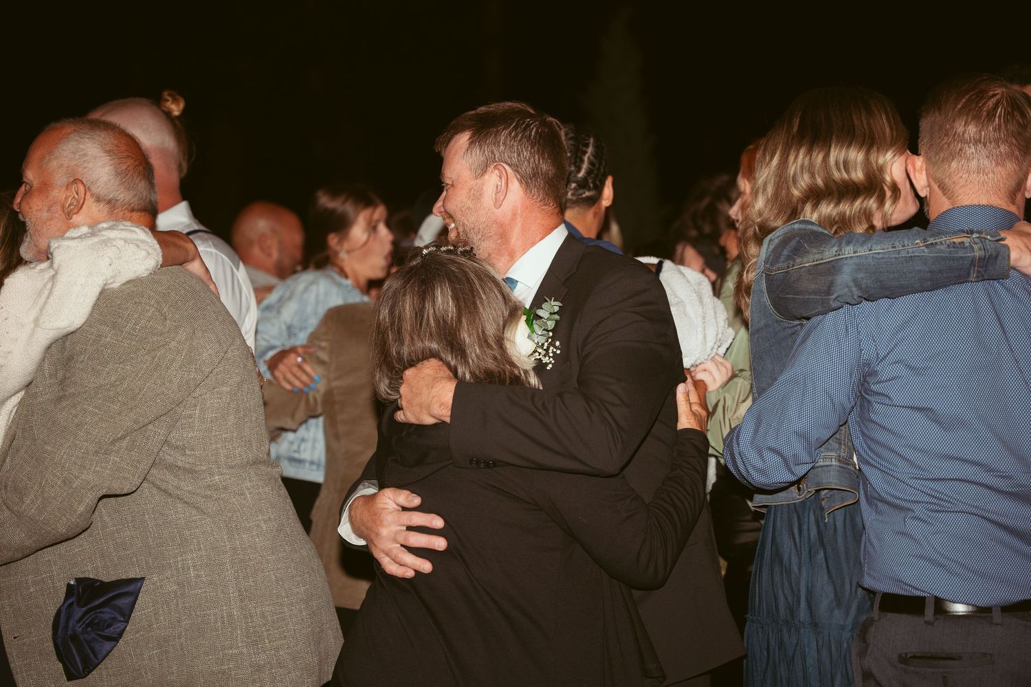 Wedding guests dance together in formal attire under dim lighting at an evening reception.