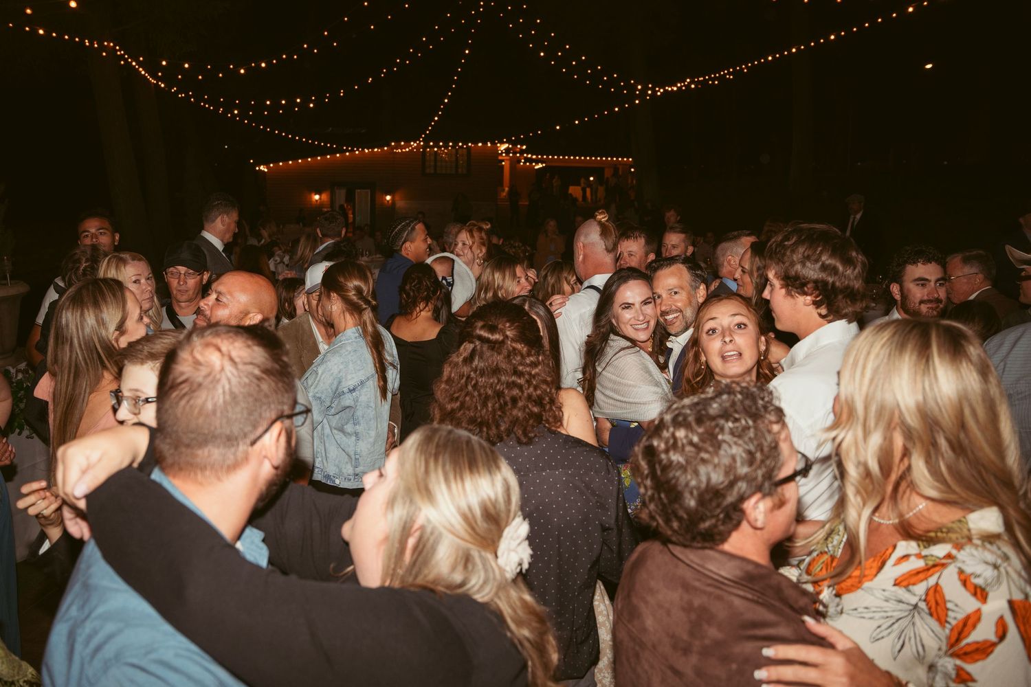 A lively crowd dances under warm string lights at an evening wedding reception celebration.