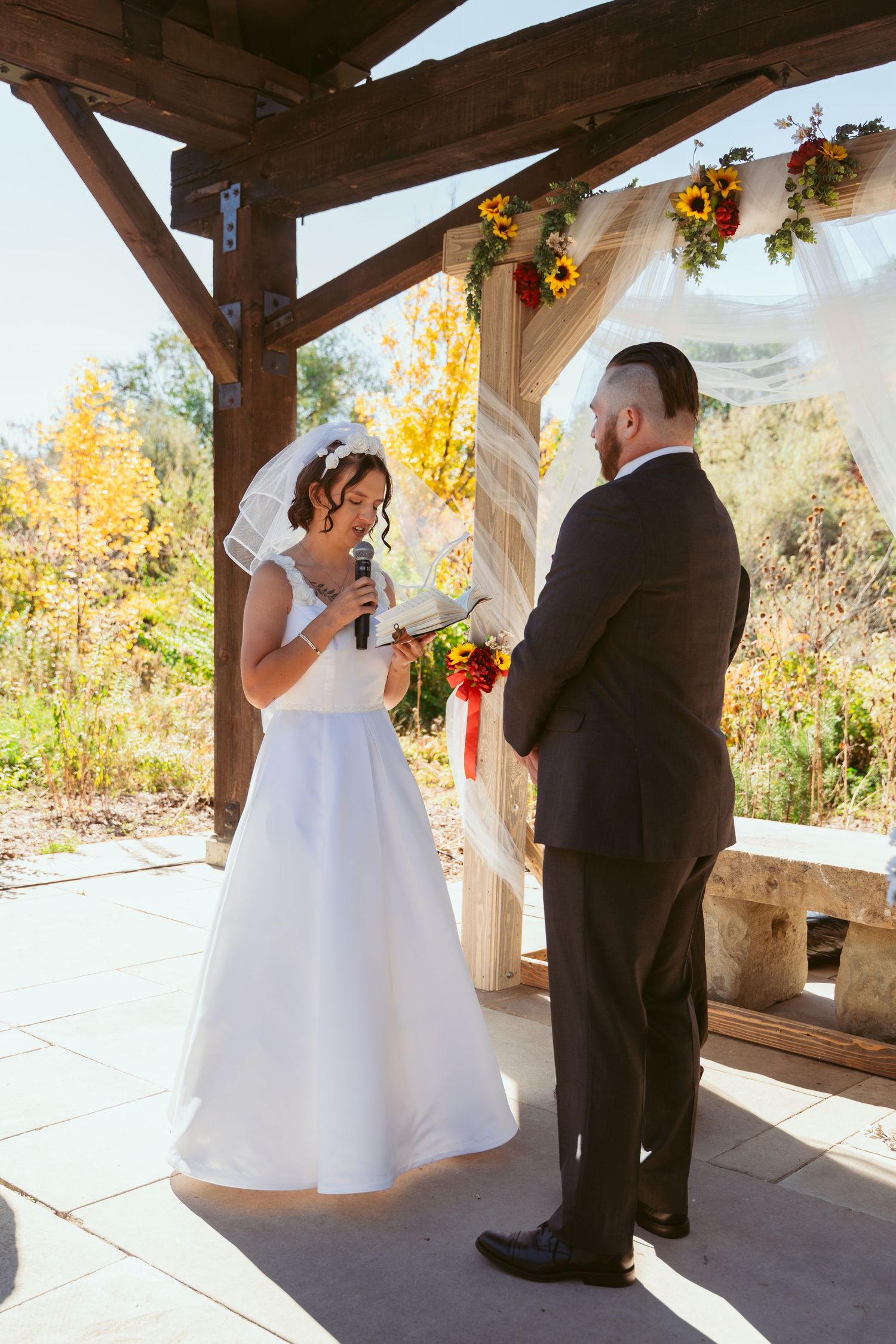 Couple exchanging vows during a fall wedding ceremony under a wooden pavilion with sunflower decorations.