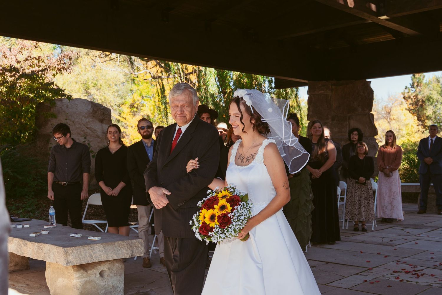 Bride holding vibrant sunflower bouquet walks down aisle at outdoor autumn wedding.