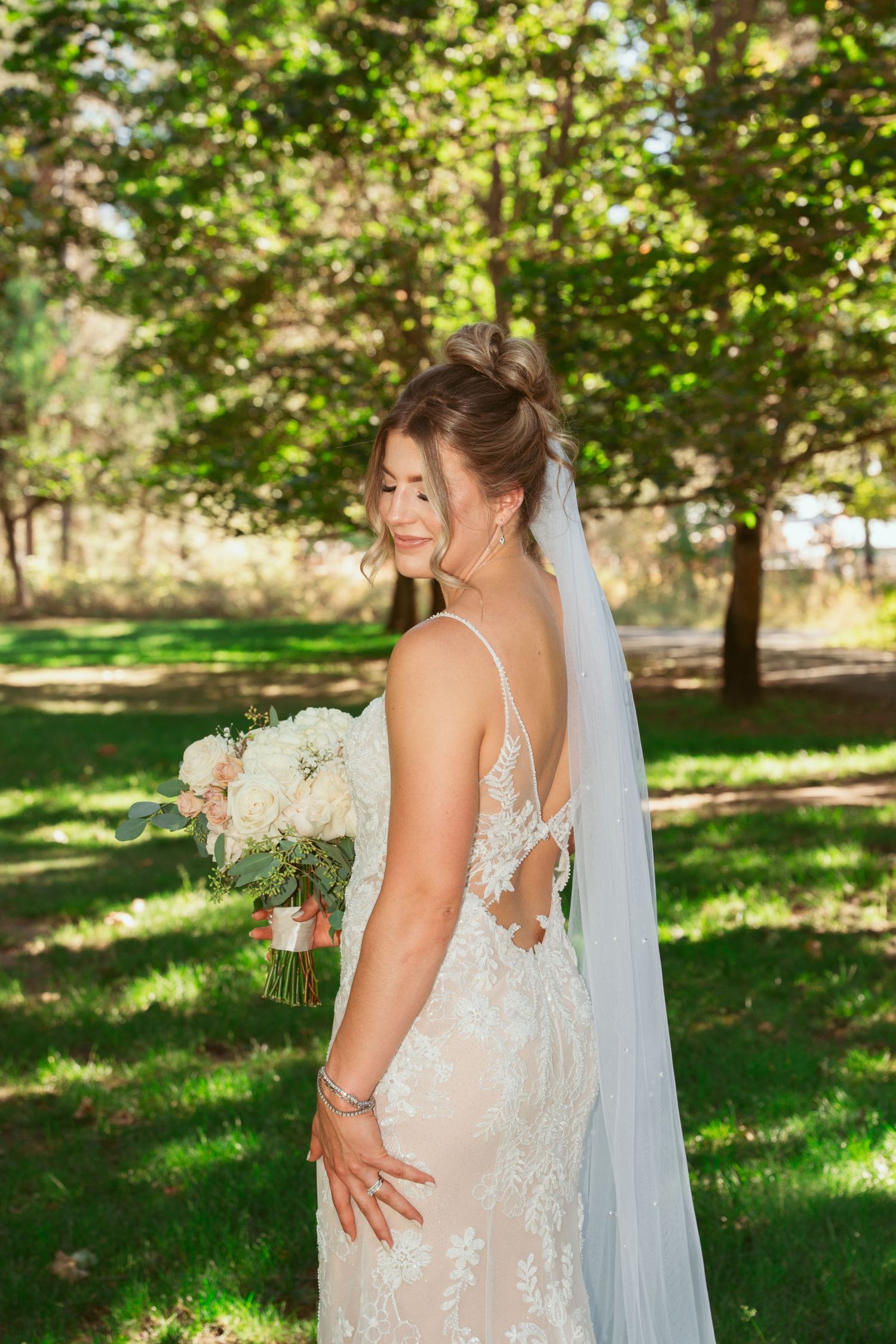 A bride in a delicate lace wedding dress poses in a sunlit garden setting.