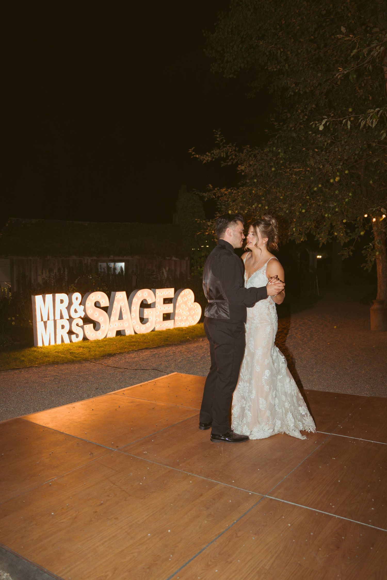 Couples dancing on an outdoor wooden dance floor with illuminated wedding signage at night.