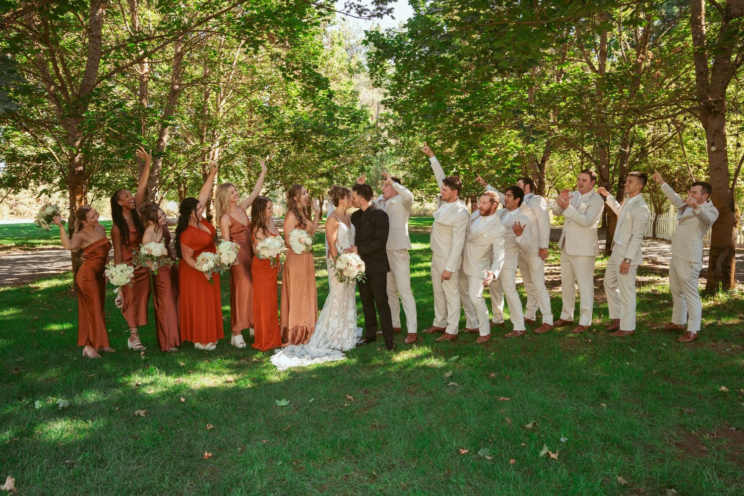 Large wedding party in white and rust-colored attire poses together on grass under tree canopy.