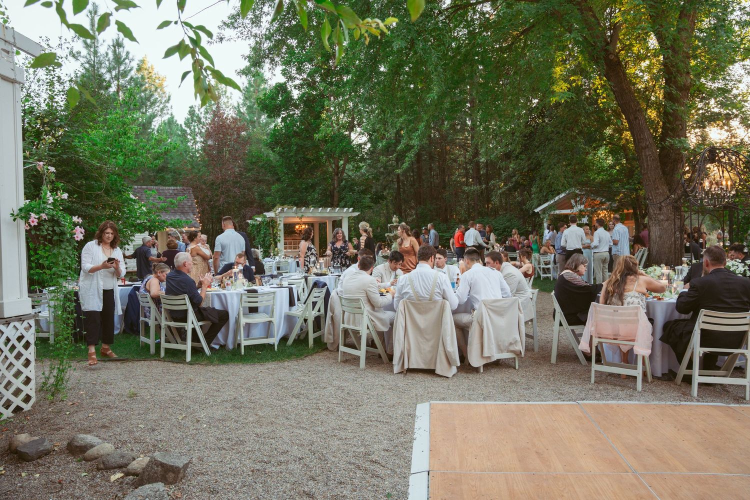 Wide view of outdoor wedding reception with white chairs and tables set up among trees at dusk.